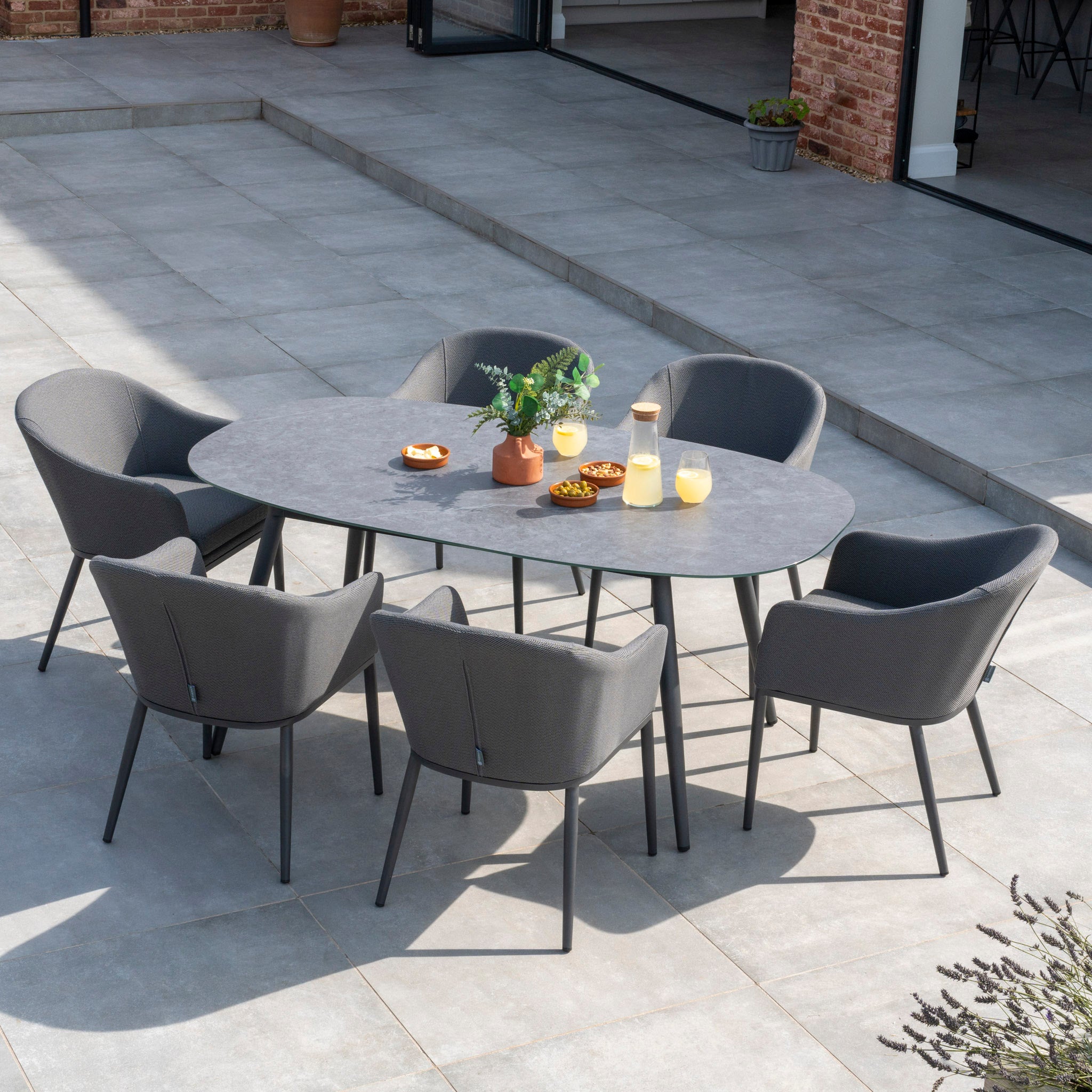 The Luna 6 Seat Outdoor Fabric Oval Ceramic Dining Set in Grey, complete with six grey chairs made from all-weather material and outdoor fabric, is beautifully arranged with drinks, snacks, and a small plant on a spacious tiled patio.