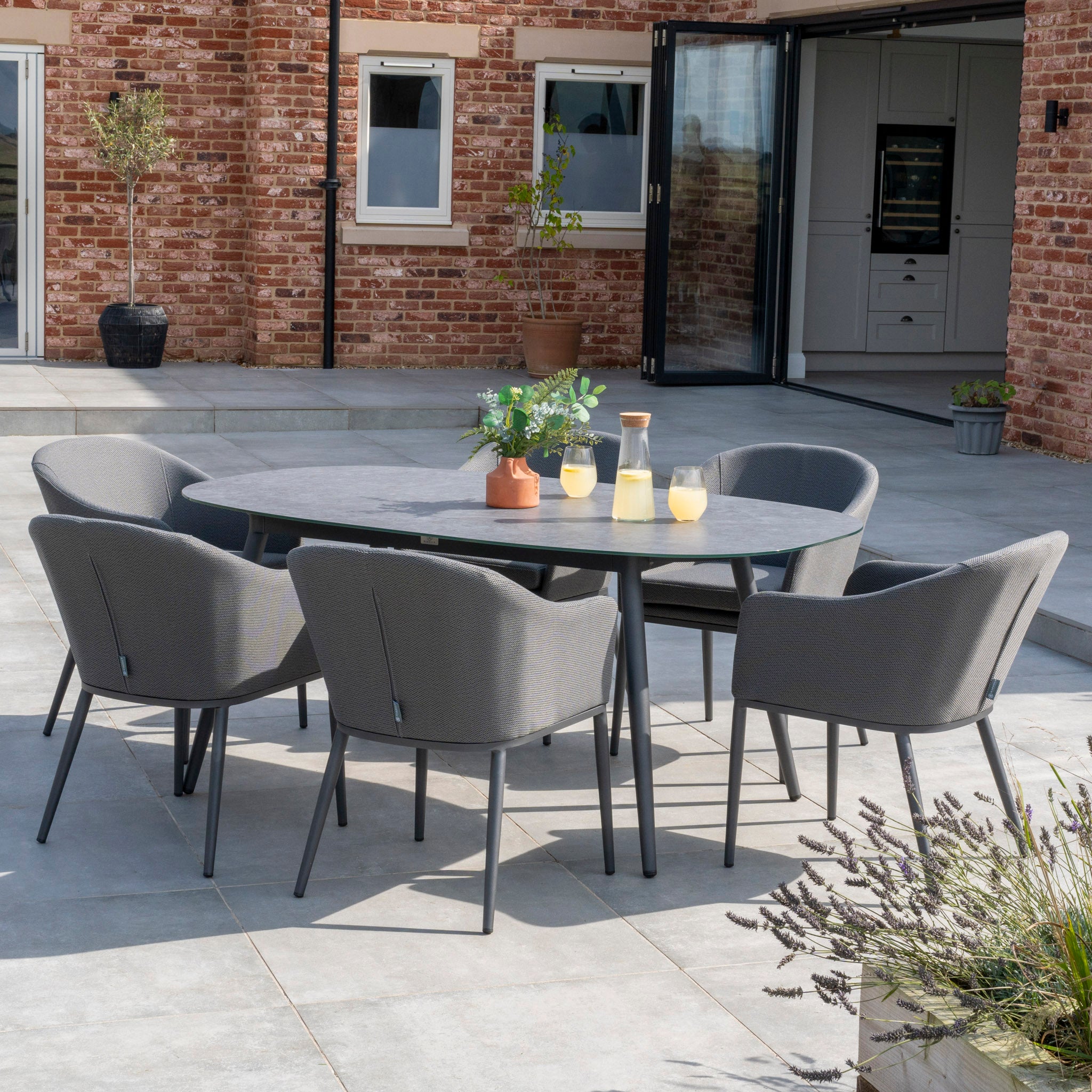 Outdoor patio setup featuring the Luna 6 Seat Outdoor Fabric Oval Ceramic Dining Set in Grey, with six gray chairs on a stone-paved area against a brick building.
