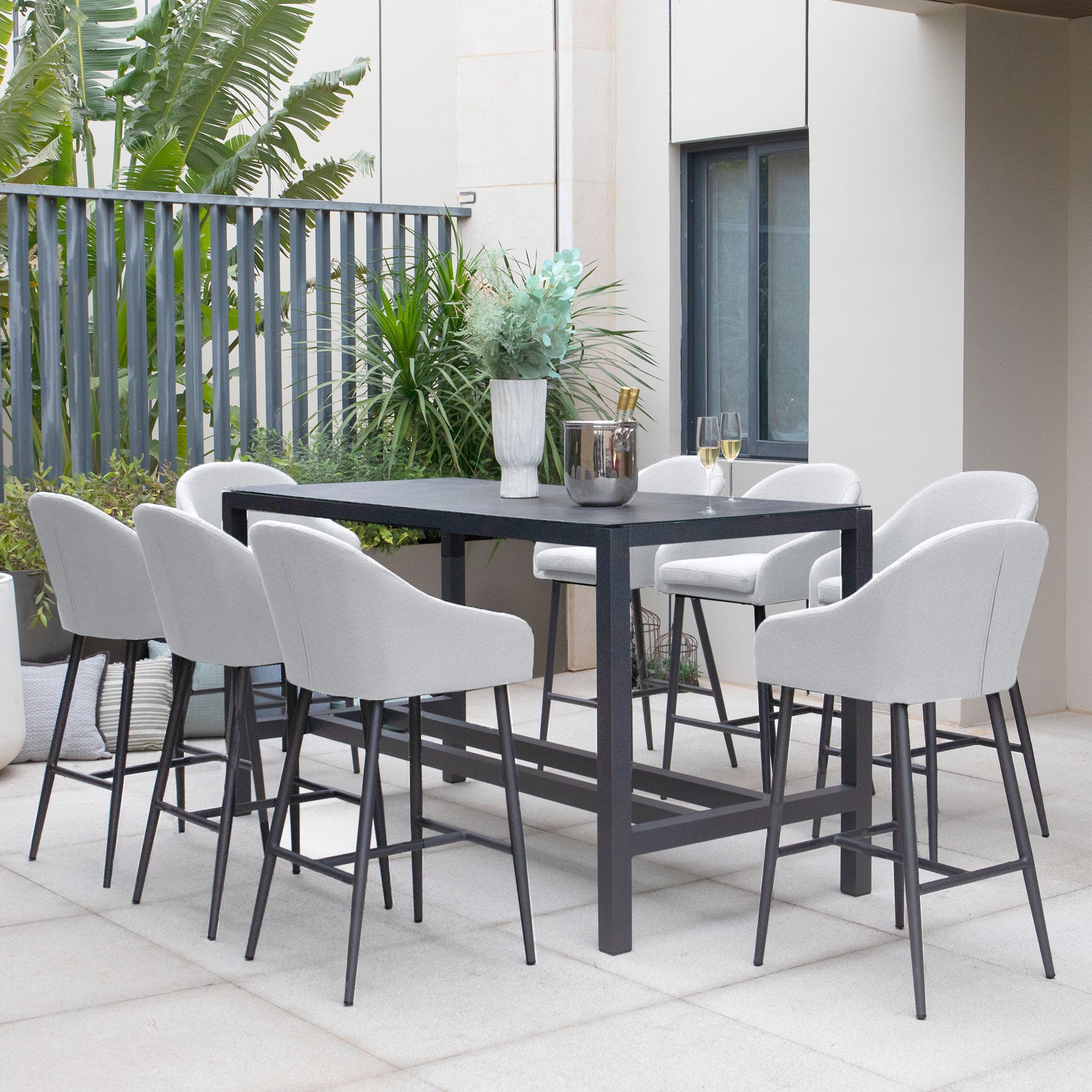 The outdoor dining area features a tall black table surrounded by eight oyster grey chairs made from Luna outdoor fabric, with greenery in the background. This stylish Luna 8 Seat Bar Set is crafted using durable all-weather material, perfect for any season.