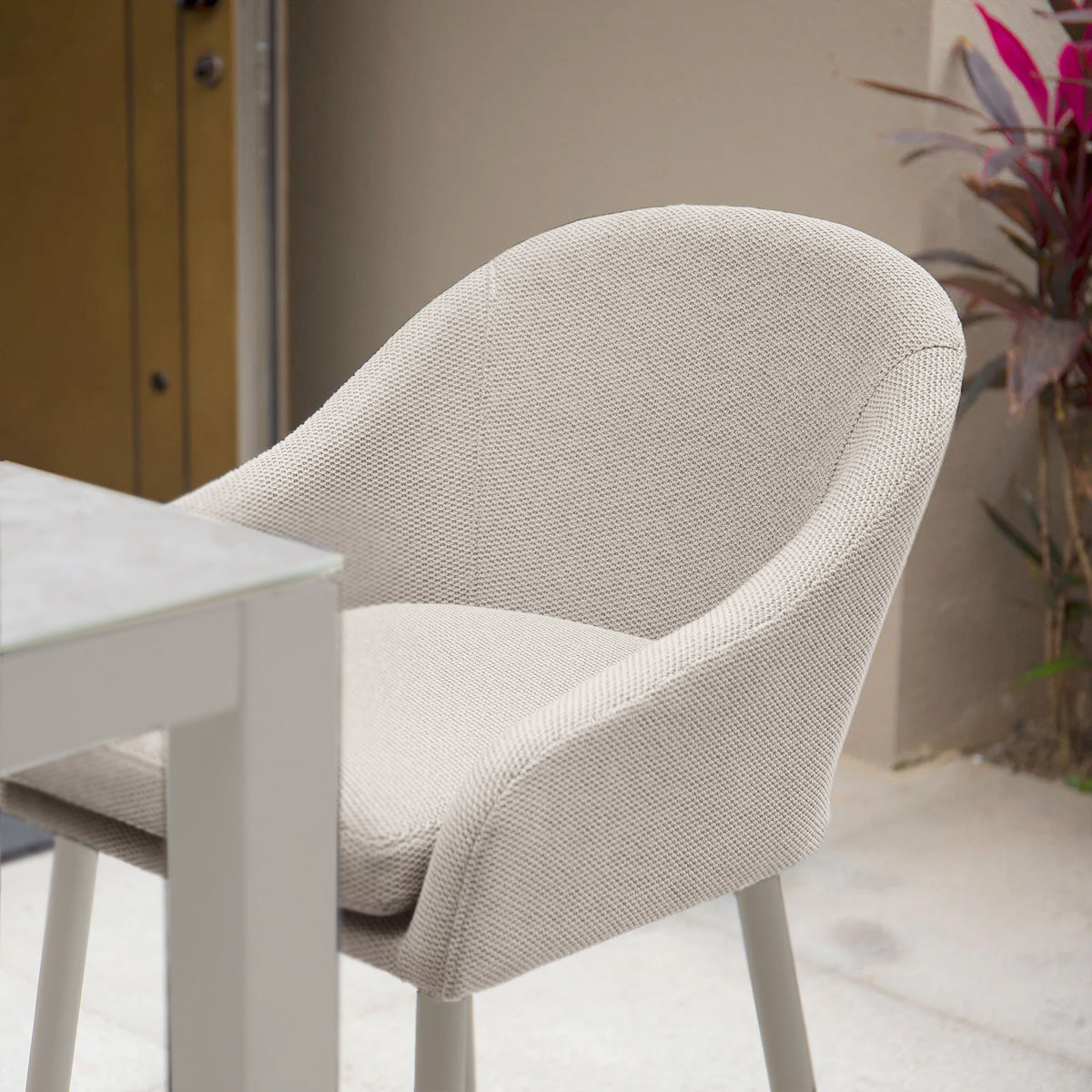 The Luna Outdoor Fabric Bar Stool in Fawn, featuring a modern design crafted from weather-resistant, stain-proof outdoor fabric, sits elegantly beside a white table, set outdoors near a lush potted plant.