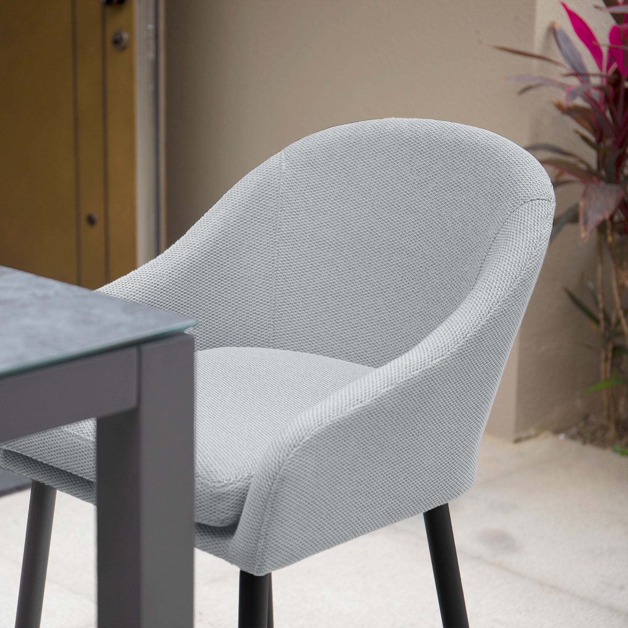 A Luna 8 Seat Bar Set in Oyster Grey features an upholstered chair with black legs, crafted from durable Luna outdoor fabric, positioned next to a dark tabletop in an outdoor setting near a beige wall and surrounded by lush plants.