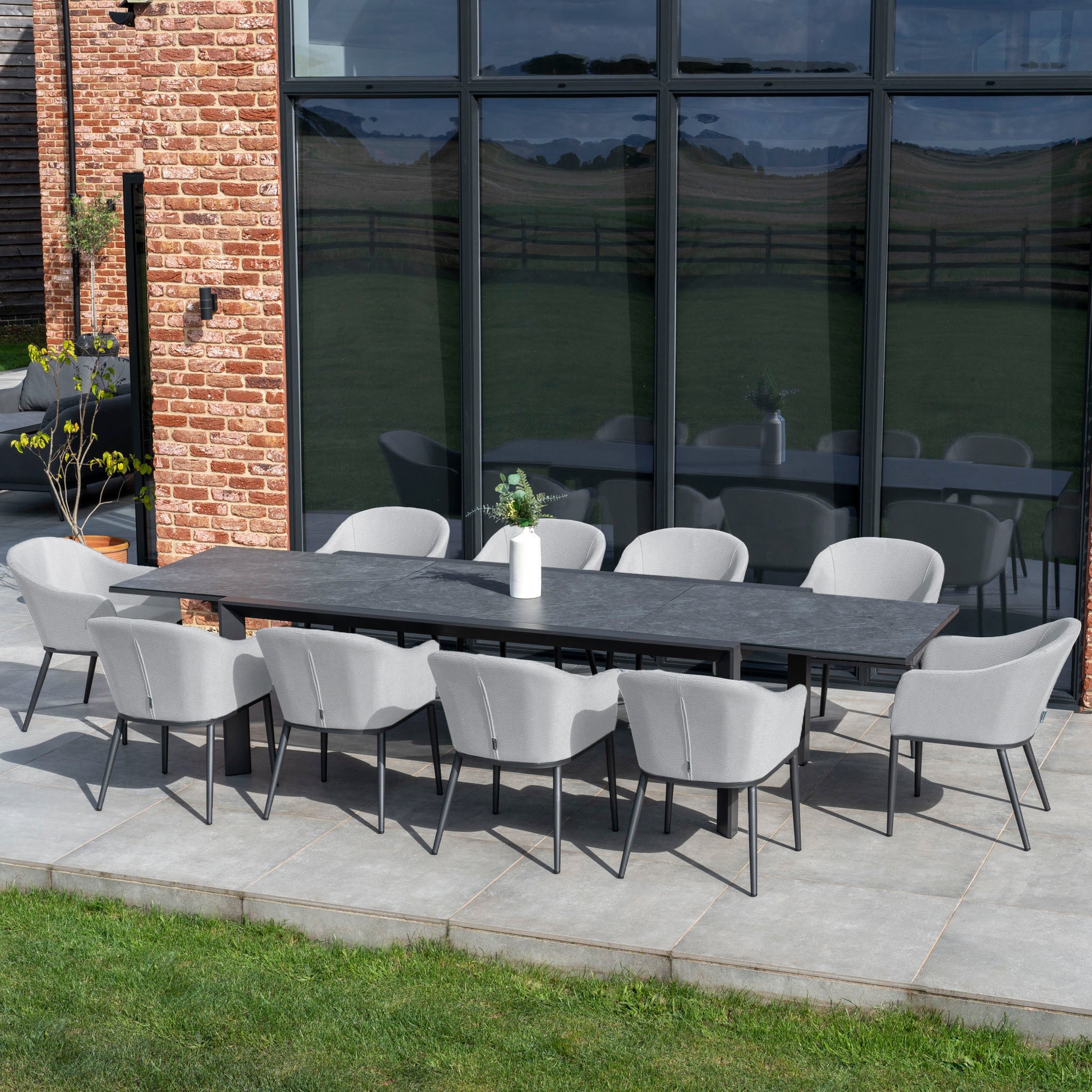 Outdoor patio featuring the Luna 8 Seat Outdoor Fabric Extending Ceramic Dining Set in Oyster Grey, positioned to face a brick wall with large windows.