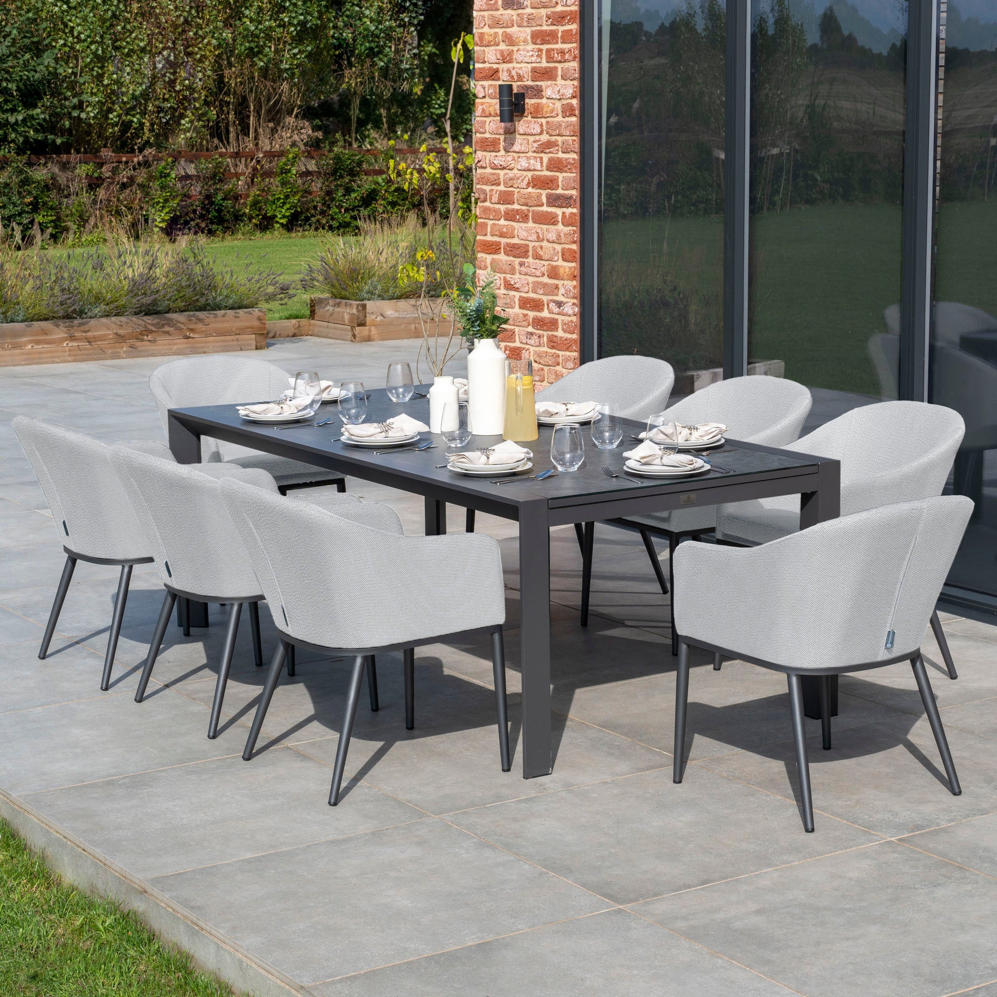 The Luna 8 Seat Outdoor Fabric Extending Ceramic Dining Set in Oyster Grey, featuring a black rectangular table and eight light gray all-weather chairs, is beautifully arranged with table settings on the patio.