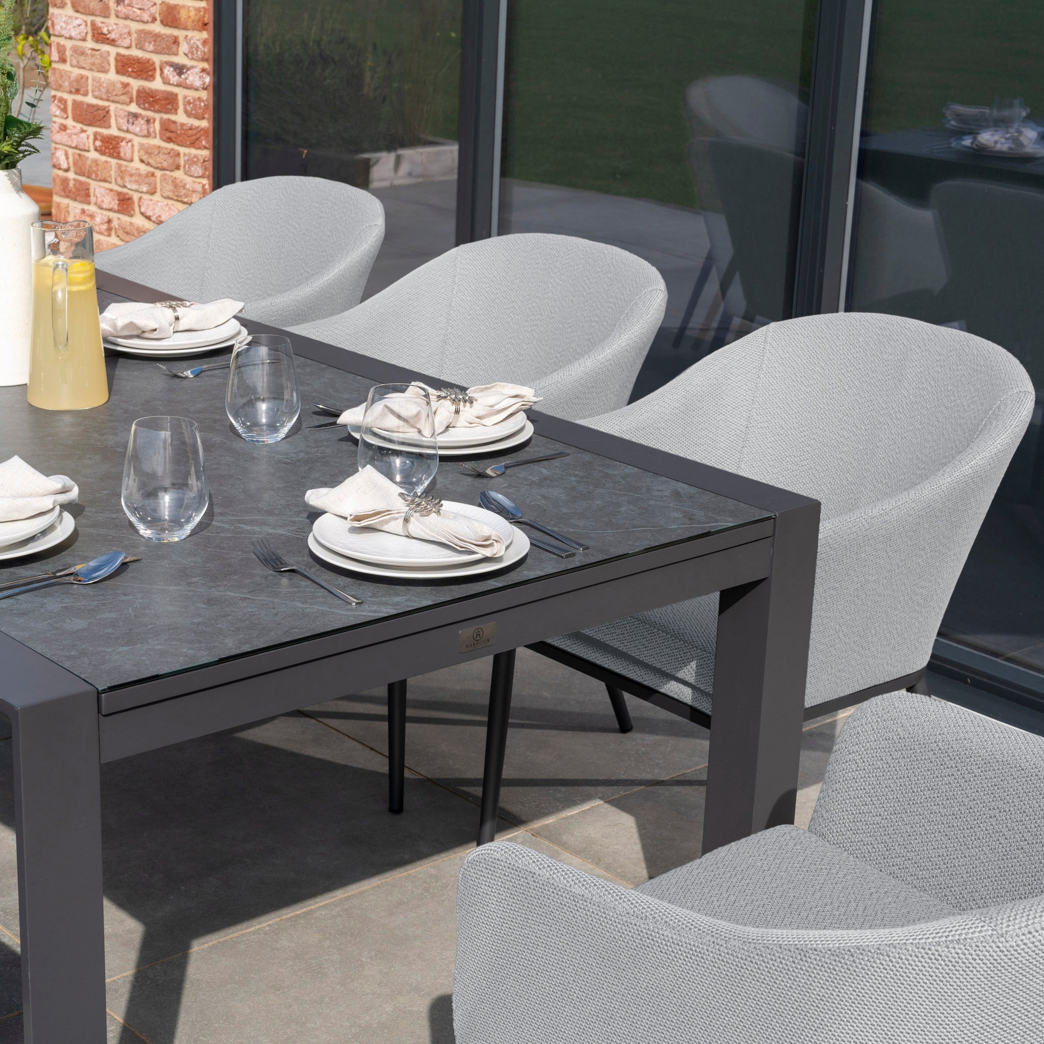 This Luna 8 Seat Outdoor Fabric Extending Ceramic Dining Set in Oyster Grey includes an outdoor dining table made from all-weather material, along with four chairs covered in durable Luna outdoor fabric, and is perfectly arranged with plates, glasses, utensils, and a yellow pitcher.