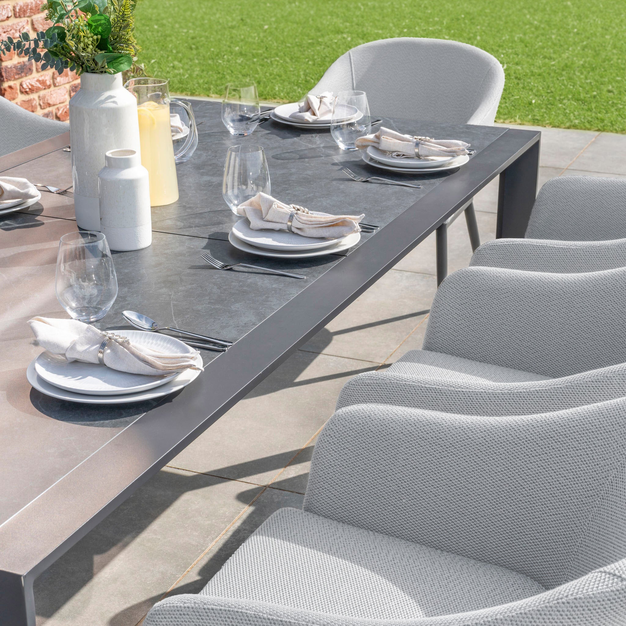 Savor al fresco meals with the Luna 8 Seat Outdoor Fabric Extending Ceramic Dining Set in Oyster Grey, complete with white plates, wine glasses, and gray cushioned chairs made from all-weather material, all set against a lush green lawn backdrop.