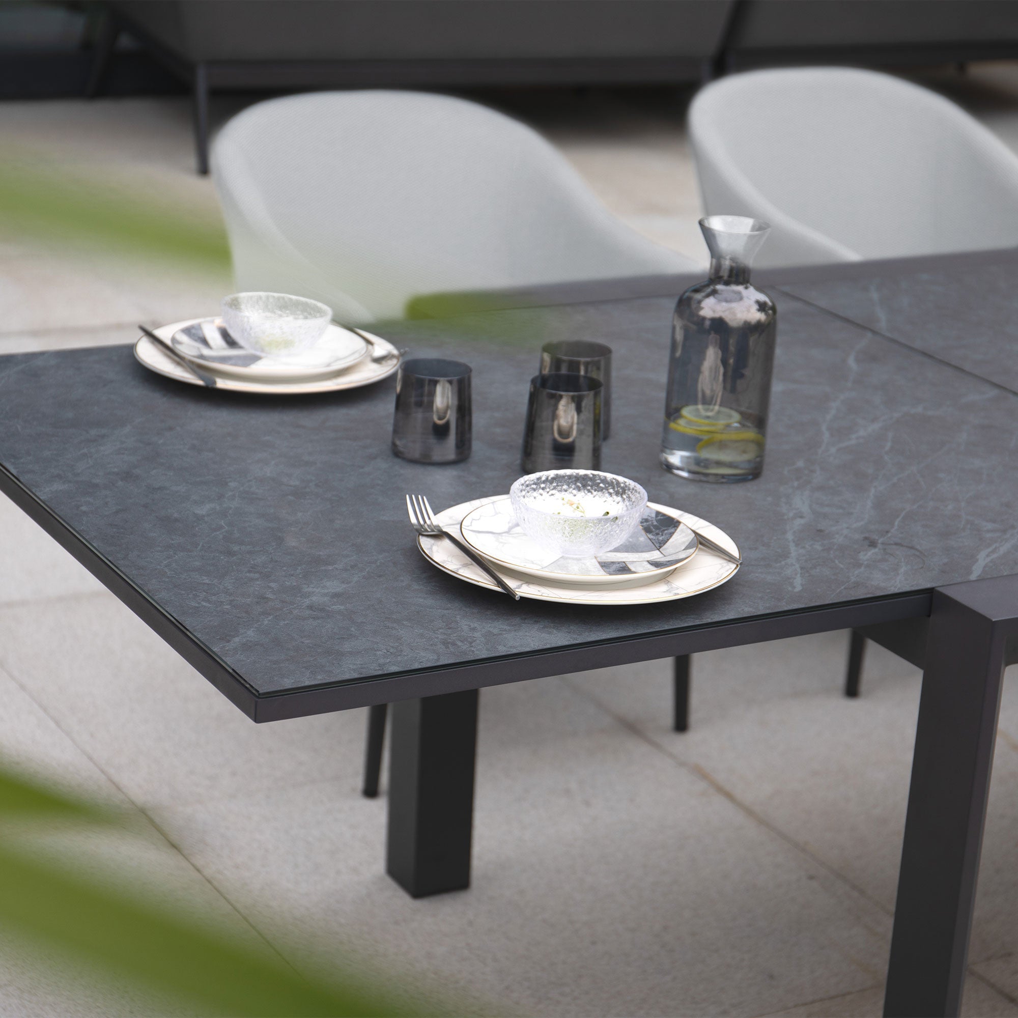 Modern outdoor dining setup for two showcasing plates, glasses, and a water carafe on a stone surface. The Luna 8 Seat Outdoor Fabric Extending Ceramic Dining Set in Oyster Grey is meticulously crafted from all-weather materials, ensuring durability through every season.