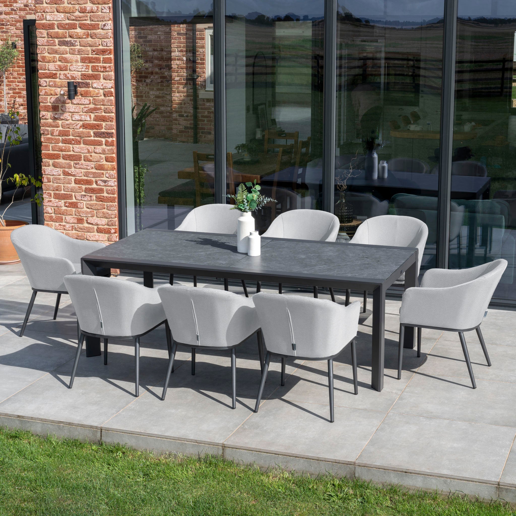 The Luna 8 Seat Outdoor Fabric Extending Ceramic Dining Set in Oyster Grey, featuring a rectangular table and eight all-weather gray chairs, is elegantly placed on the patio next to a brick wall and large windows.