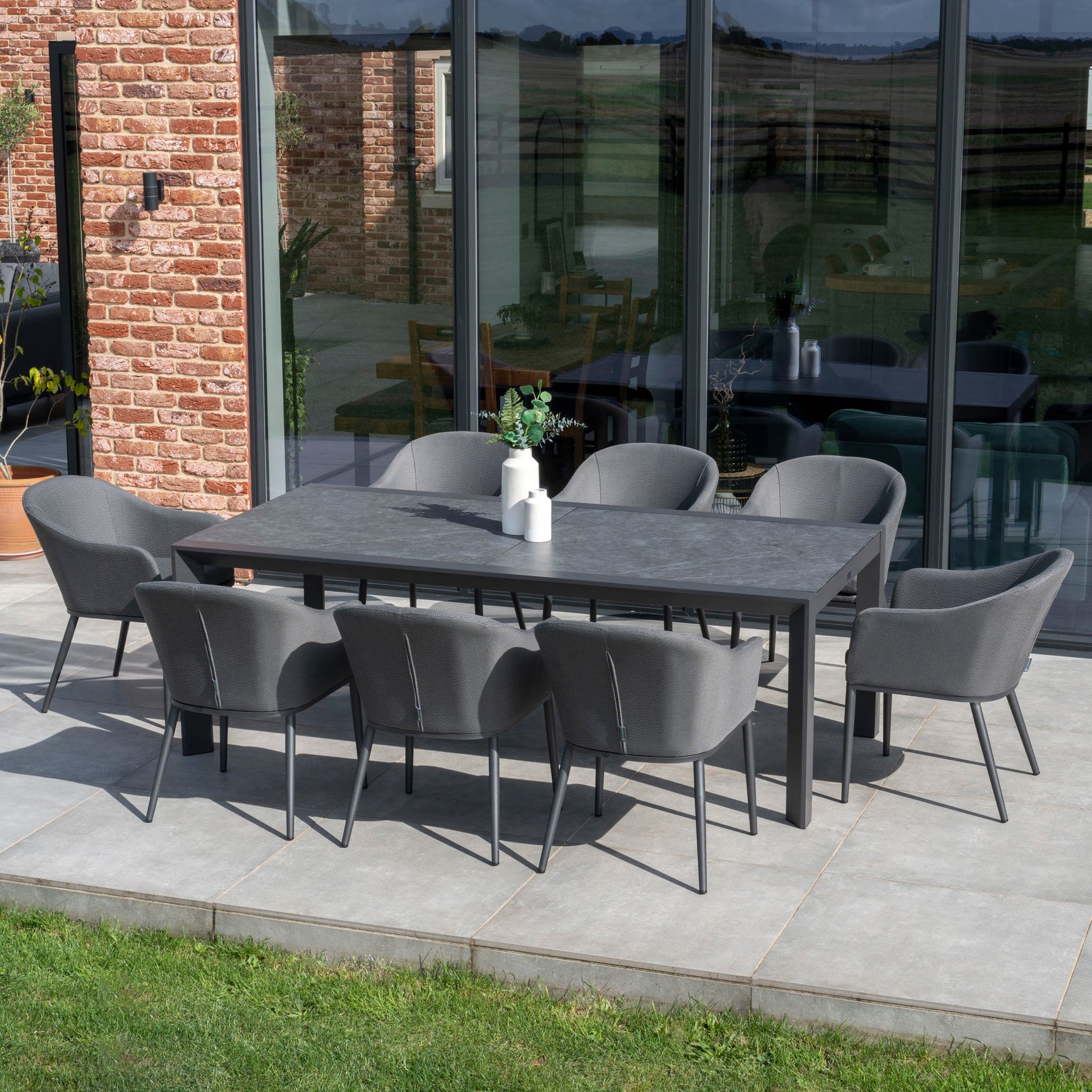 The Luna 8 Seat Outdoor Fabric Extending Ceramic Dining Set in Grey, with eight chairs around a rectangular table, is elegantly arranged on a concrete terrace by a brick house.