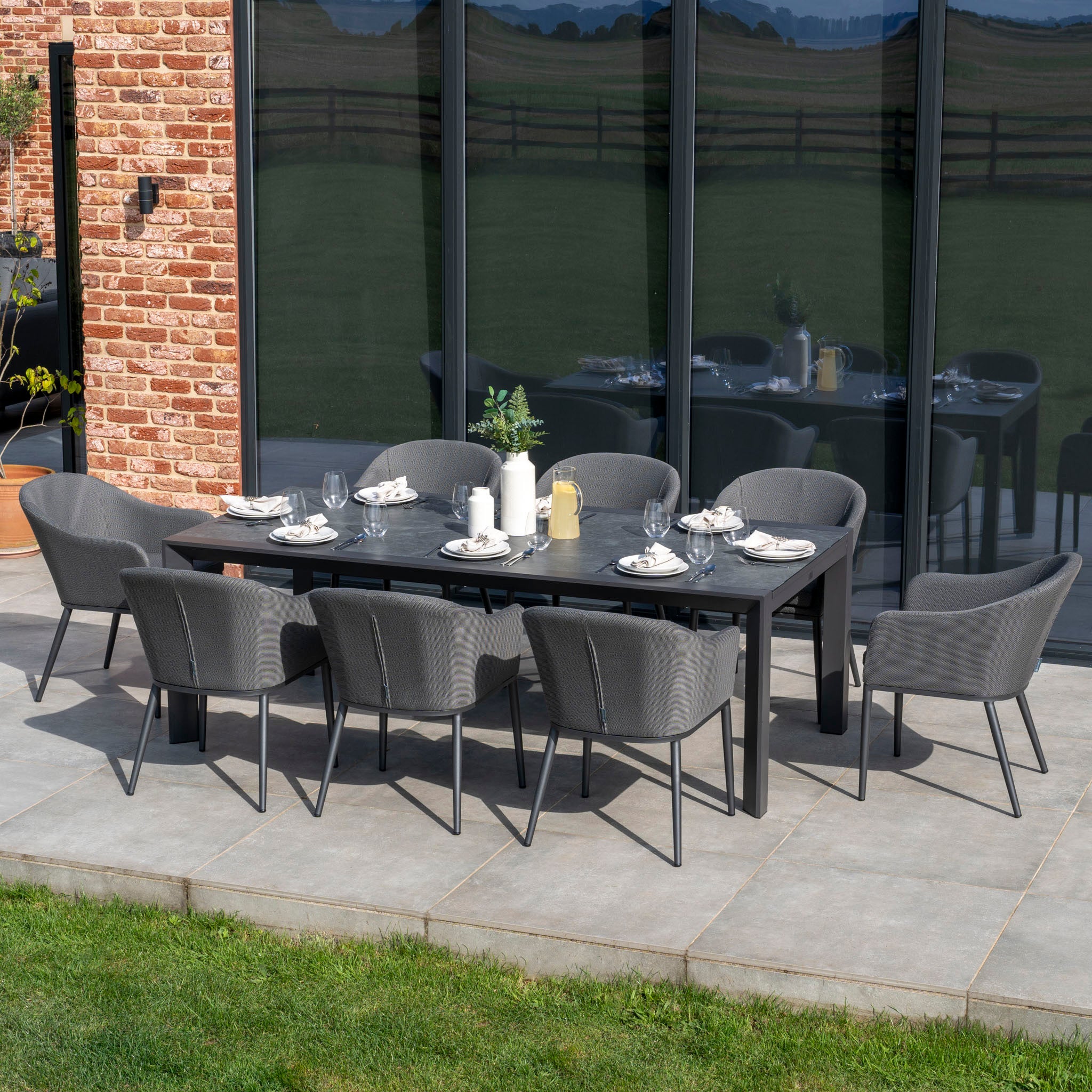 Luna 8 Seat Outdoor Fabric Extending Ceramic Dining Set in Grey, featuring a modern rectangular table, eight grey chairs, and complete table settings, showcased on a patio with lush green grass.