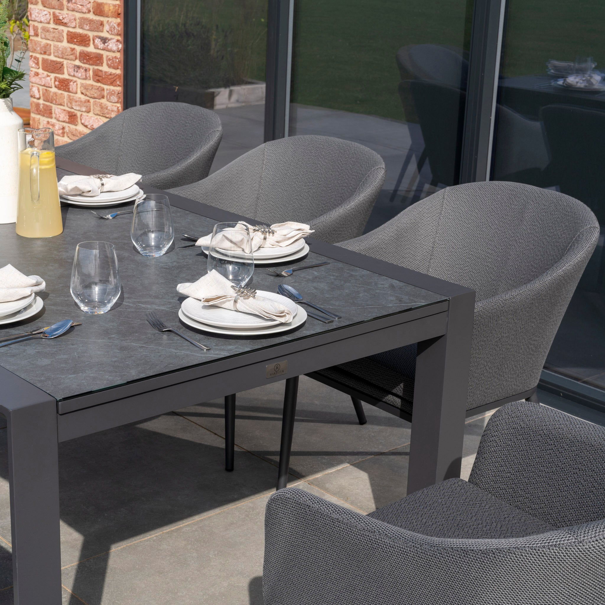 The Luna 8 Seat Outdoor Fabric Extending Ceramic Dining Set in Grey includes stain-proof chairs with cushioned seats and a dark rectangular table, complete with plates, glasses, and a pitcher.