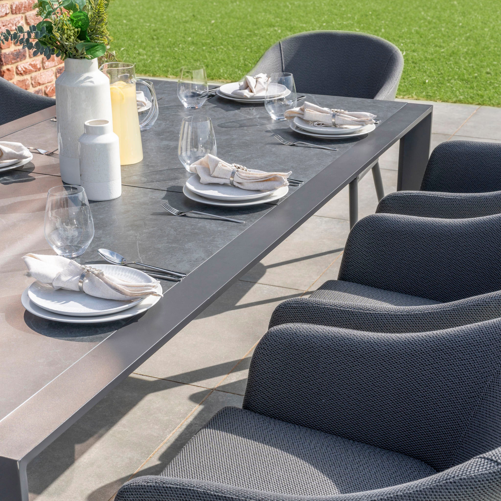 The Luna 8 Seat Outdoor Fabric Extending Ceramic Dining Set in Grey includes plates, napkins, wine glasses, and modern gray chairs for your patio. This rust-proof dining set is ideal for all weather conditions. Enjoy your meals with the green lawn as a backdrop.
