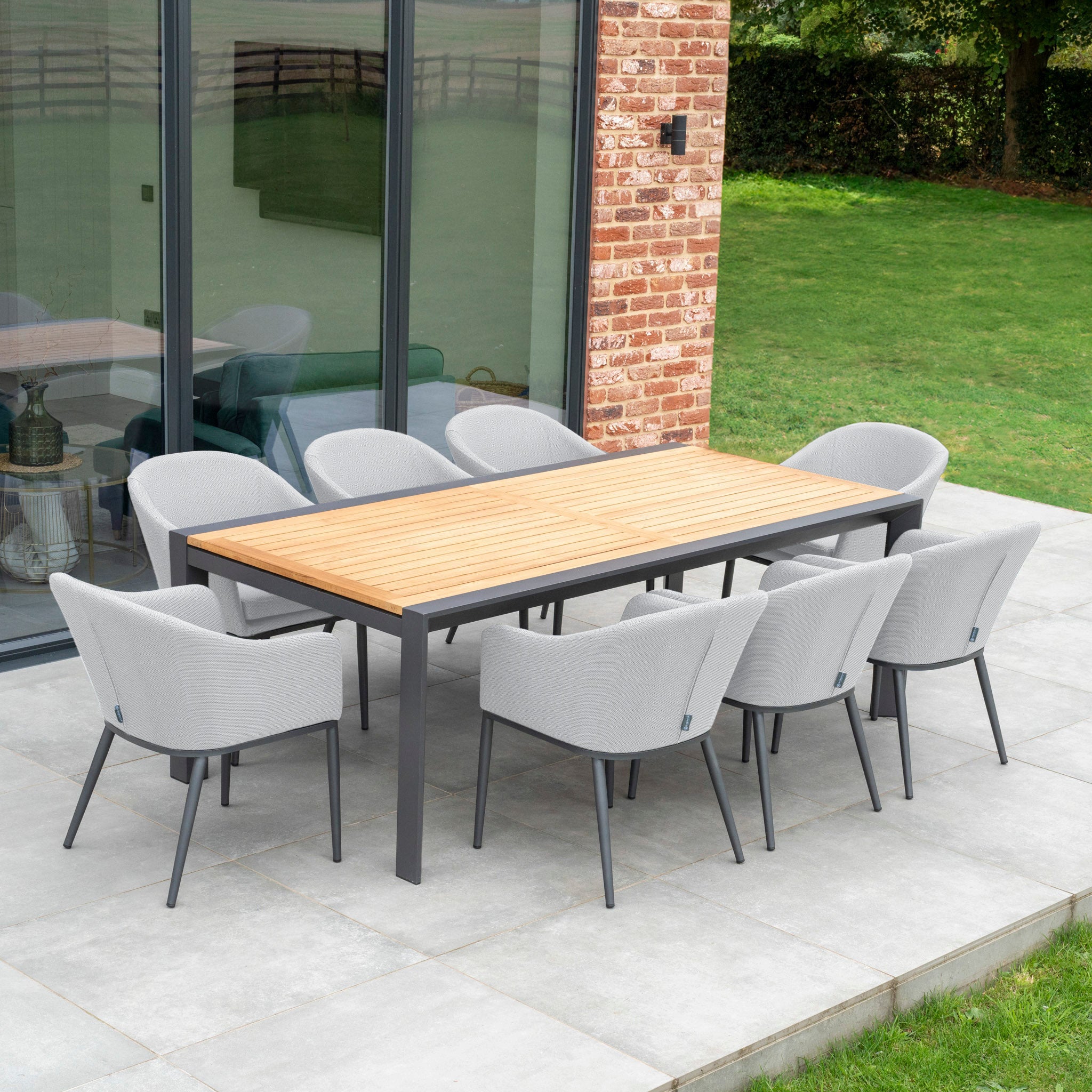 The Luna 8 Seat Outdoor Fabric Extending Teak Dining Set in Oyster Grey is elegantly arranged on a patio next to a brick house and green lawn, featuring a wooden table and eight grey cushioned chairs designed for maintenance-free care.