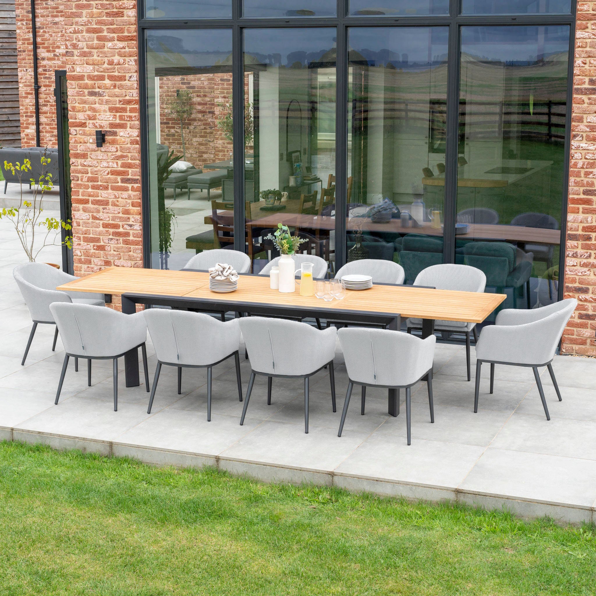 The Luna 8 Seat Outdoor Fabric Extending Teak Dining Set in Oyster Grey graces the patio near a brick wall and large glass windows reflecting the horizon. Its maintenance-free outdoor fabric ensures durability and comfort for every gathering.
