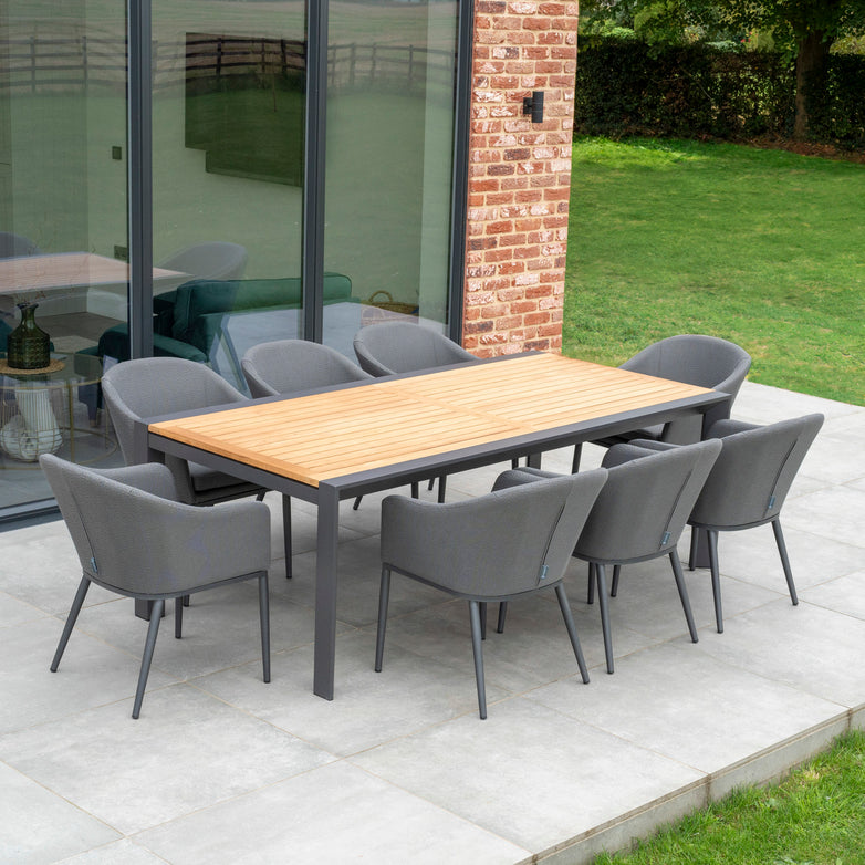 A patio featuring the Luna 8 Seat Outdoor Fabric Extending Teak Dining Set in Grey, which boasts stain-proof and rainproof properties, accompanied by gray cushioned chairs on a concrete surface adjacent to a glass wall and brick exterior.