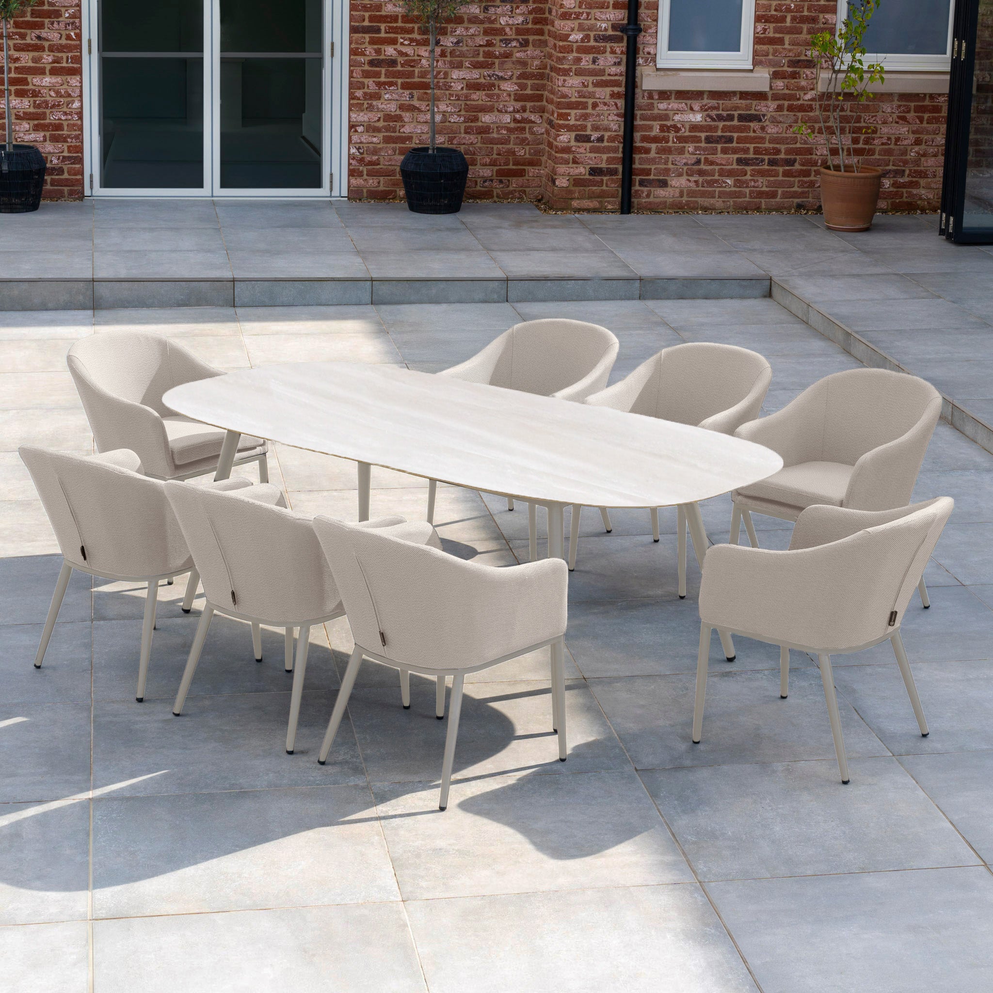 The Luna 8 Seat Outdoor Fabric Oval Ceramic Dining Set in Fawn features eight beige chairs and an oval table, perfect for patios with stone flooring, brick walls, and potted plants.
