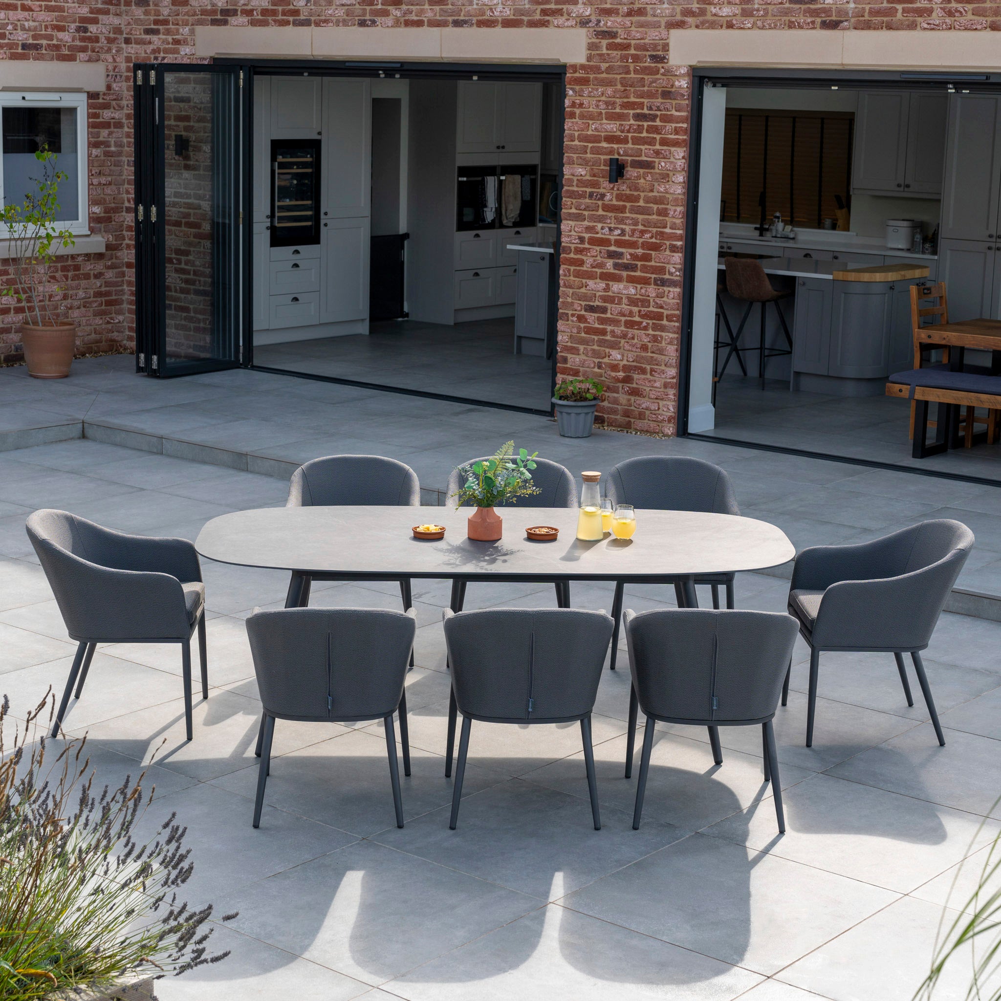 Luna 8 Seat Outdoor Fabric Oval Ceramic Dining Set in Grey. This elegant gray table and eight matching chairs are made with all-weather materials, perfect for your patio outside a brick house with open sliding doors.