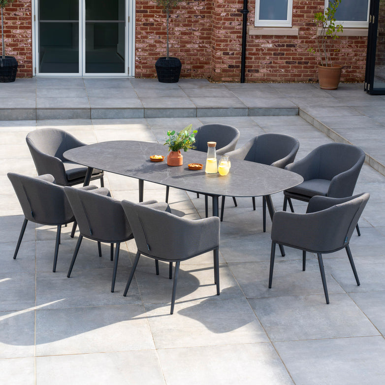 Outdoor patio with a Luna 8 Seat Outdoor Fabric Oval Ceramic Dining Set in Grey, featuring cushioned chairs made from all-weather material, and a few potted plants in the background.