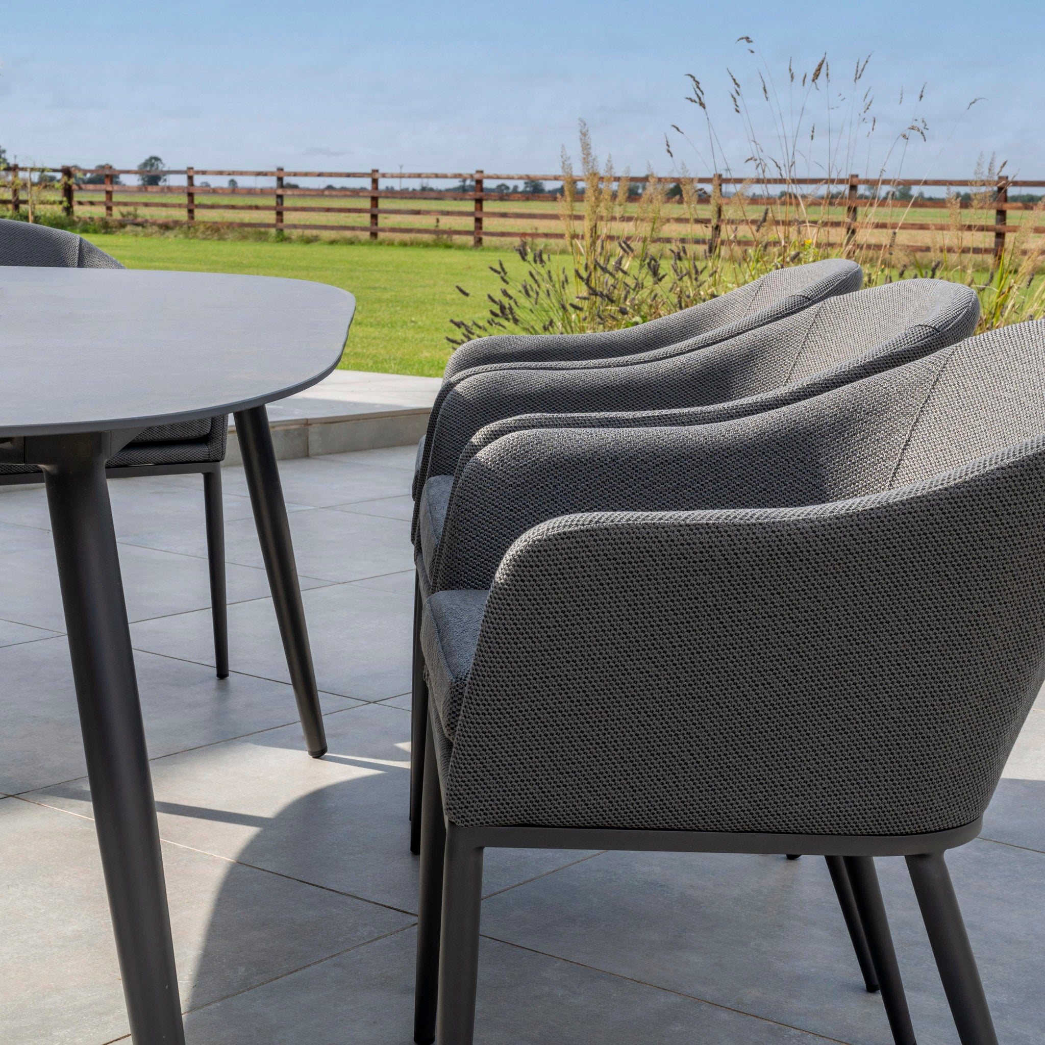 The Luna 8 Seat Outdoor Fabric Oval Ceramic Dining Set in Grey is arranged on a tiled outdoor area, overlooking a grassy field and wooden fence.