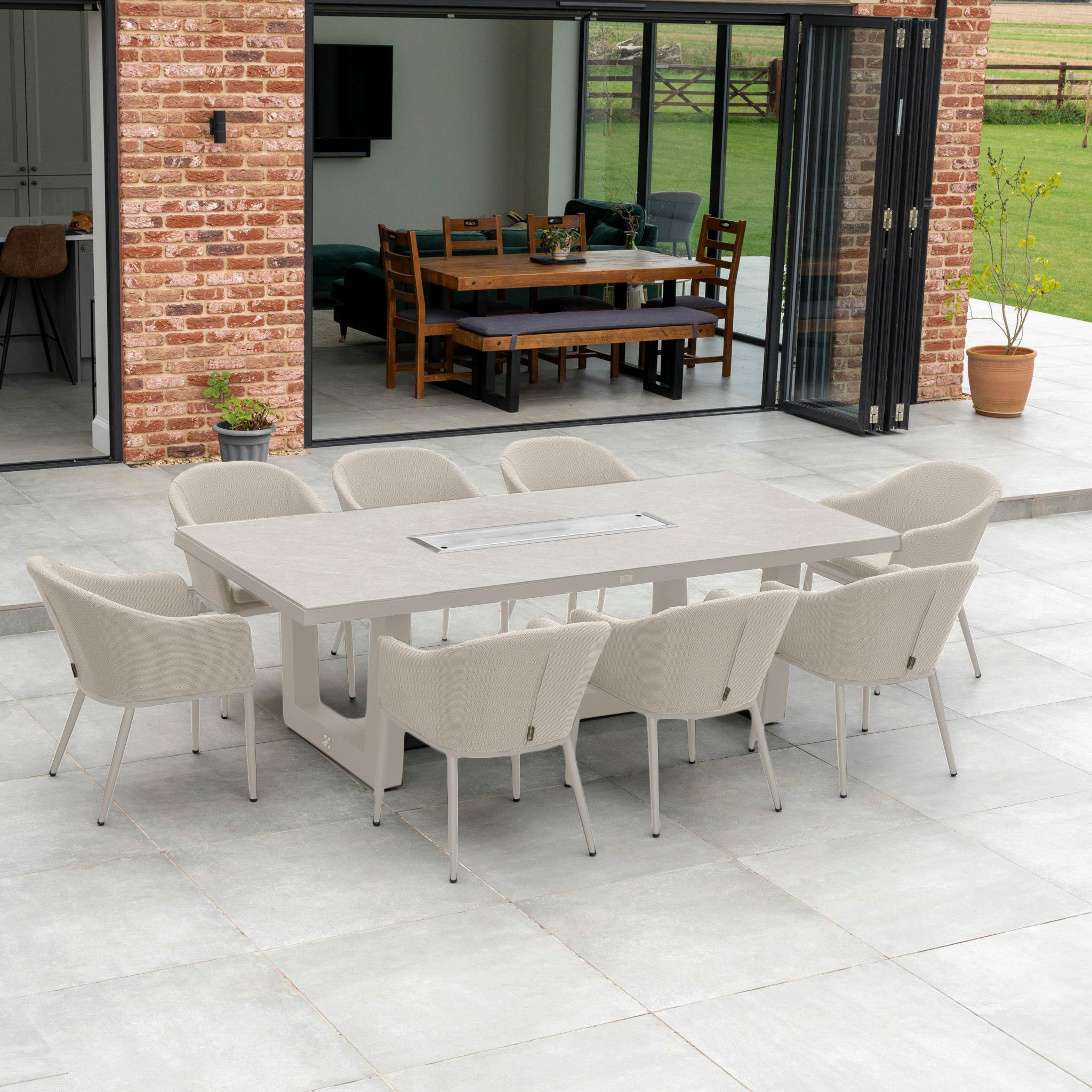 Set the stage with the Luna 8 Seat Outdoor Fabric Rectangular Ceramic Firepit Dining Set in Fawn, showcasing its elegant gray chairs and table on a tiled patio, beautifully set against a brick wall with open doors in the background.