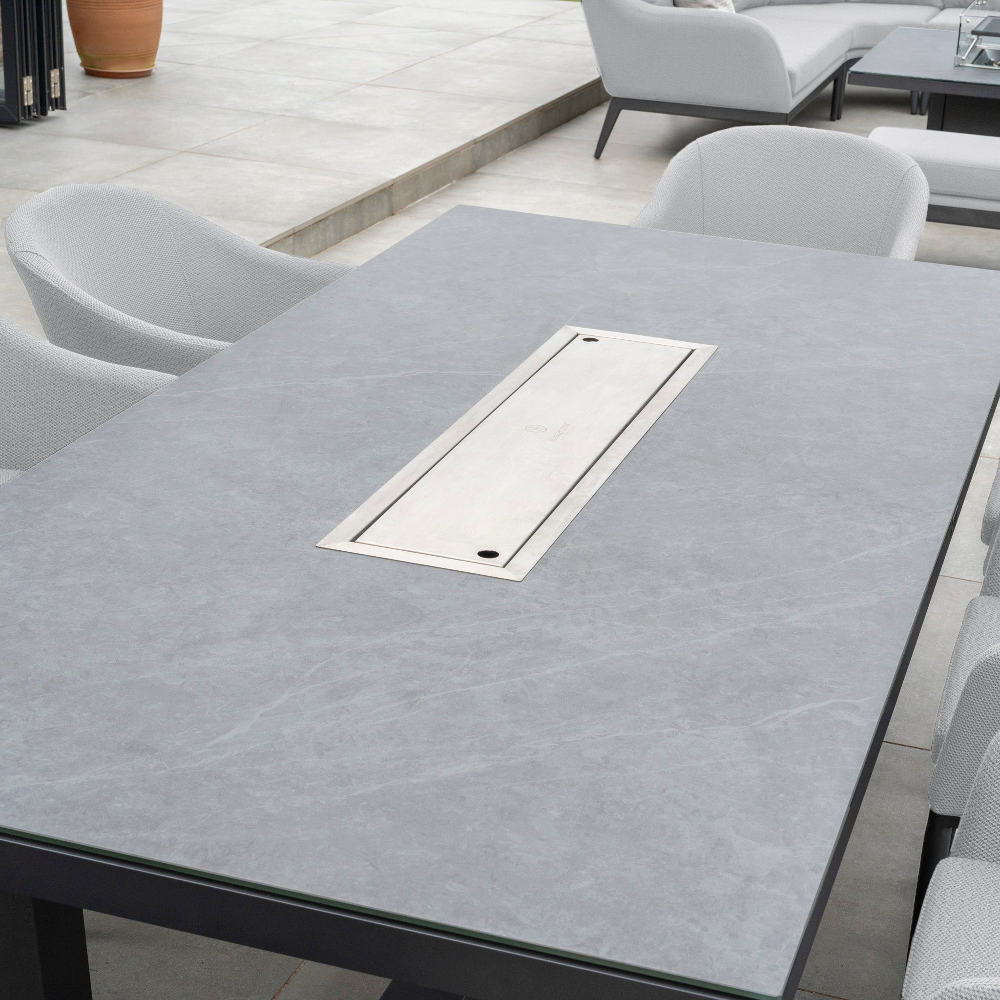 Modern outdoor dining table with a sleek, rectangular inset and cushioned gray chairs on a tiled patio, featuring the Luna 8 Seat Outdoor Fabric Rectangular Ceramic Firepit Dining Set in Oyster Grey from Harbour Lifestyle.