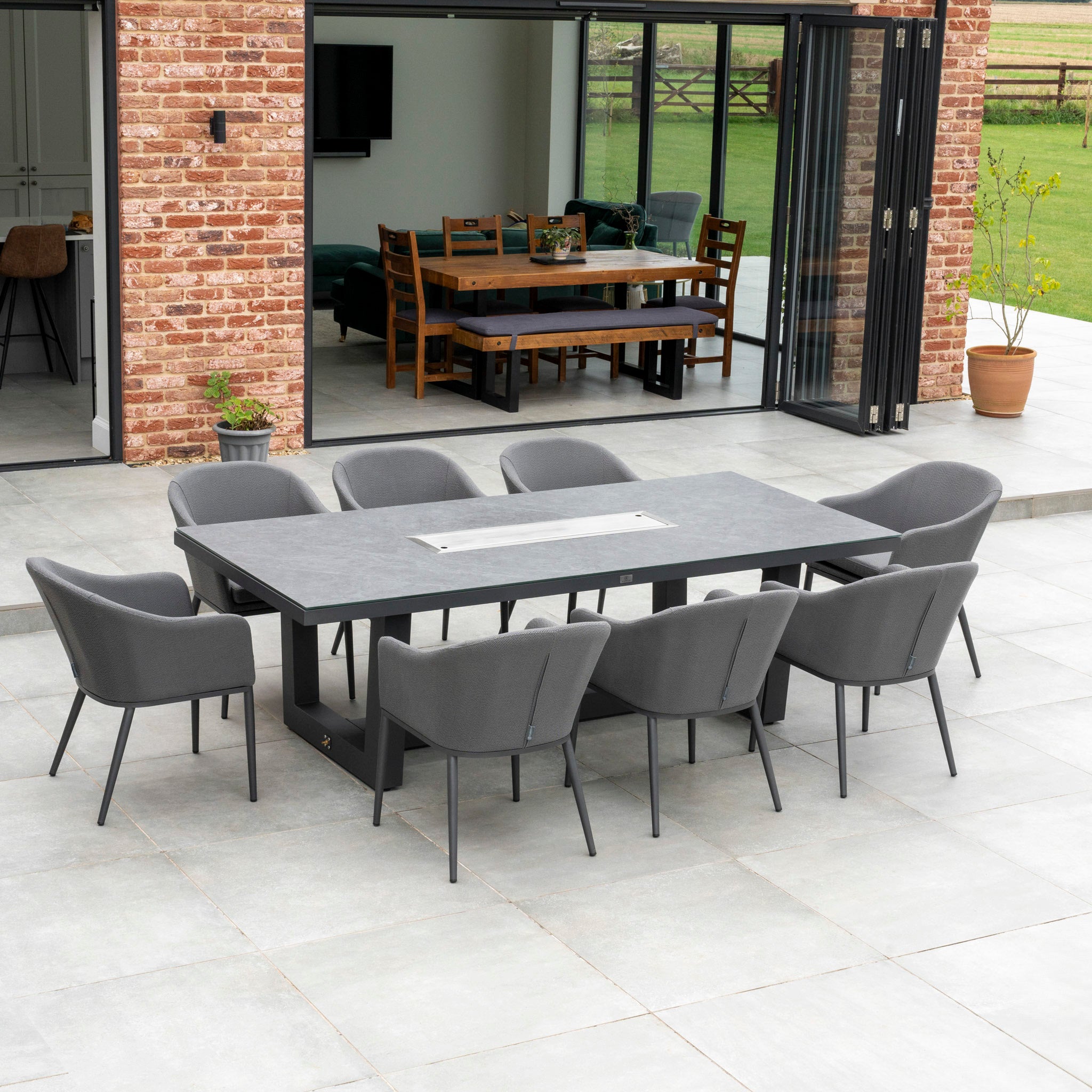 A Luna 8 Seat Outdoor Fabric Rectangular Ceramic Firepit Dining Set in Grey, featuring all-weather chairs, adorns a spacious tiled terrace near an open brick house.