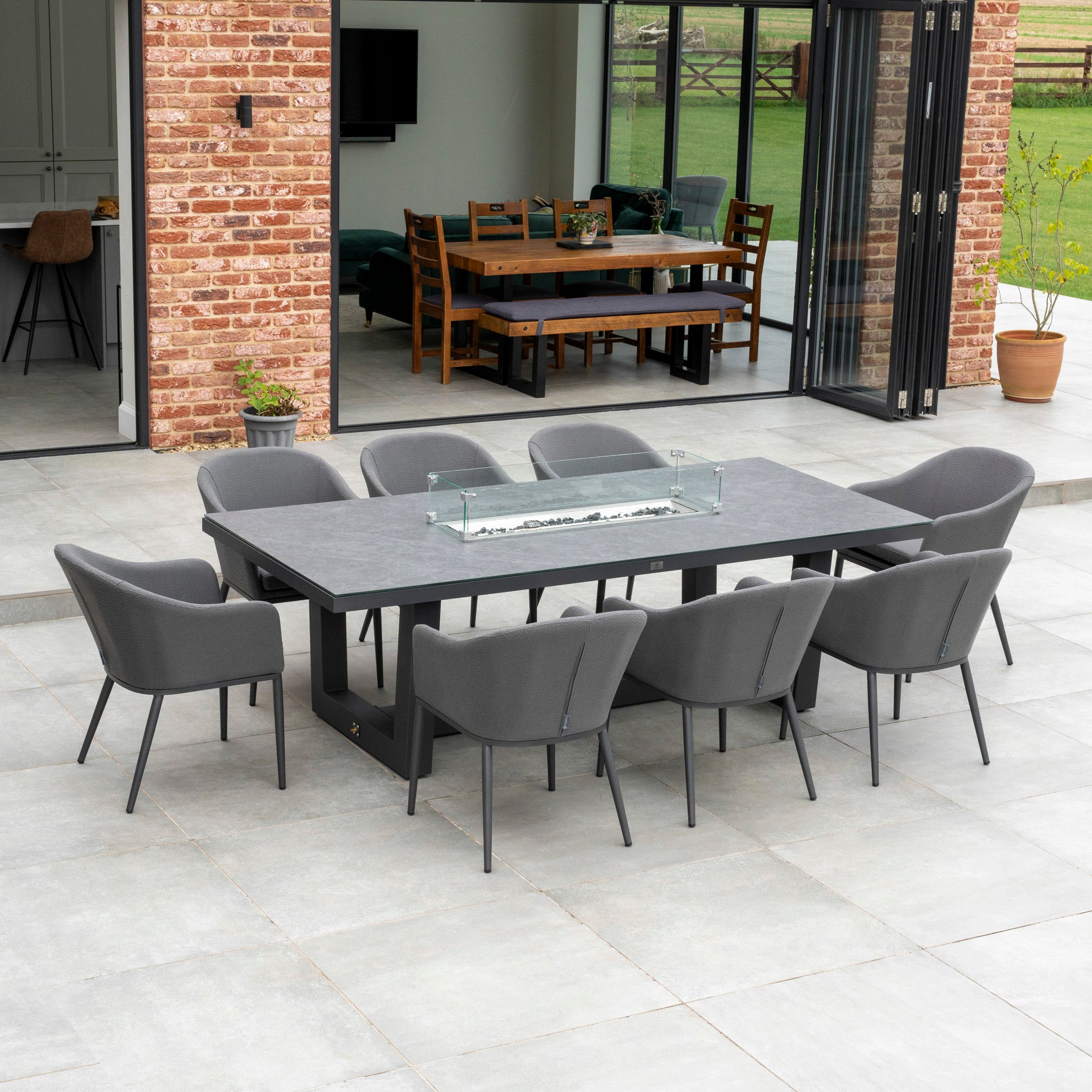 Outdoor dining area with the Luna 8 Seat Outdoor Fabric Rectangular Ceramic Firepit Dining Set in Grey. The long table is surrounded by eight gray cushioned chairs, near a building with brick walls and glass doors. The chairs are crafted from all-weather material, making this set perfect for year-round enjoyment.