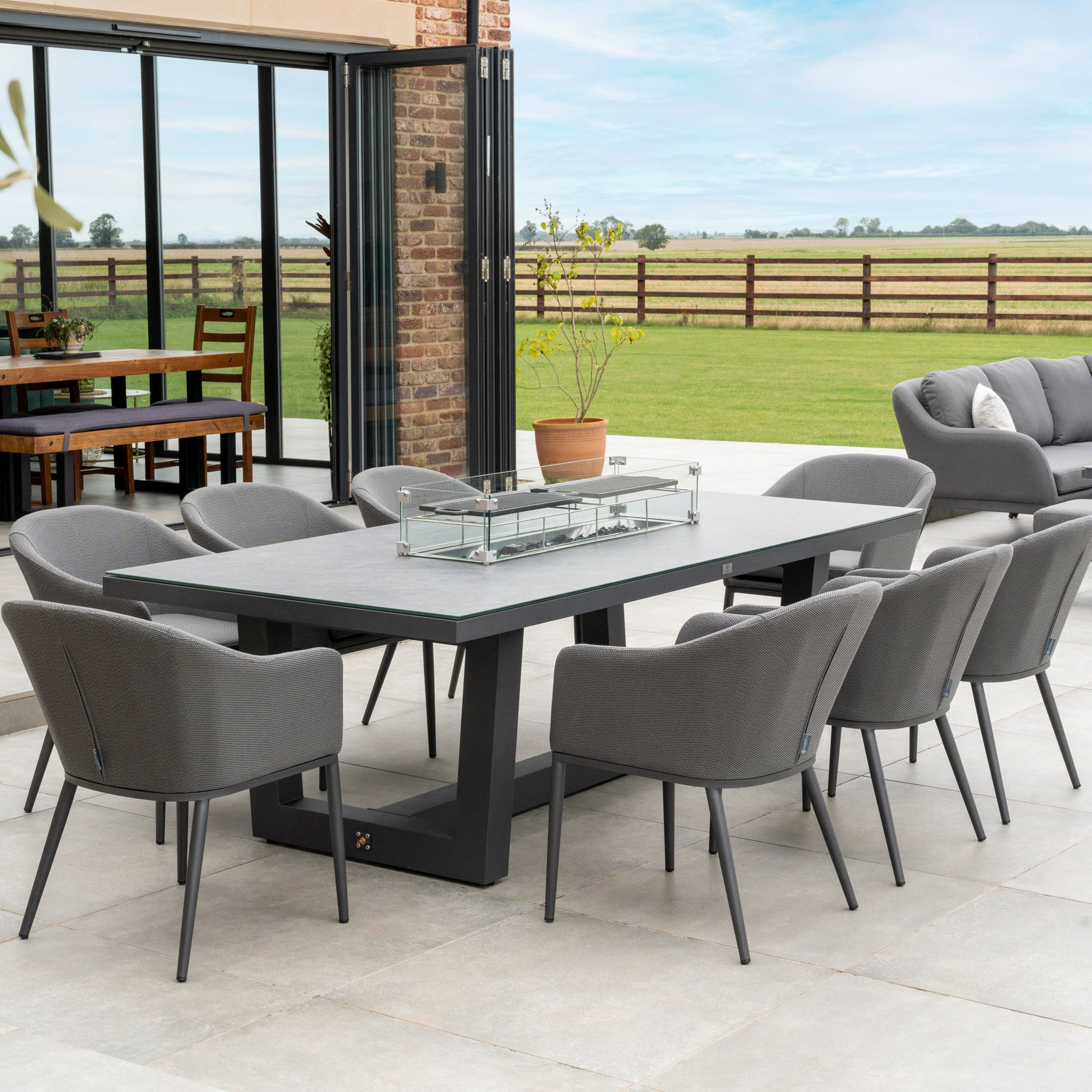 Modern outdoor dining area featuring the Luna 8 Seat Outdoor Fabric Rectangular Ceramic Firepit Dining Set in Grey, with a large rectangular table and eight gray chairs crafted from durable all-weather material. Complemented by Luna outdoor fabric cushions, the setup is situated against the backdrop of a fenced field.