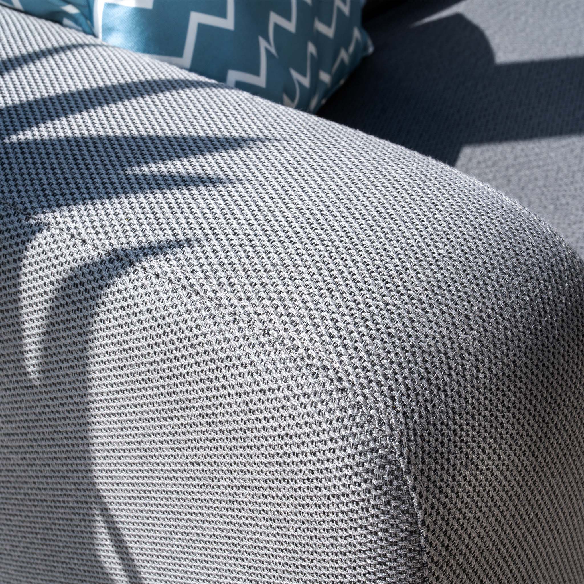 A close-up of the Luna Outdoor Fabric Large Corner Group Set with Firepit Coffee Table in Grey showcases sunlight casting shadows on the textured couch, accented by a blue pillow with white zigzag patterns.