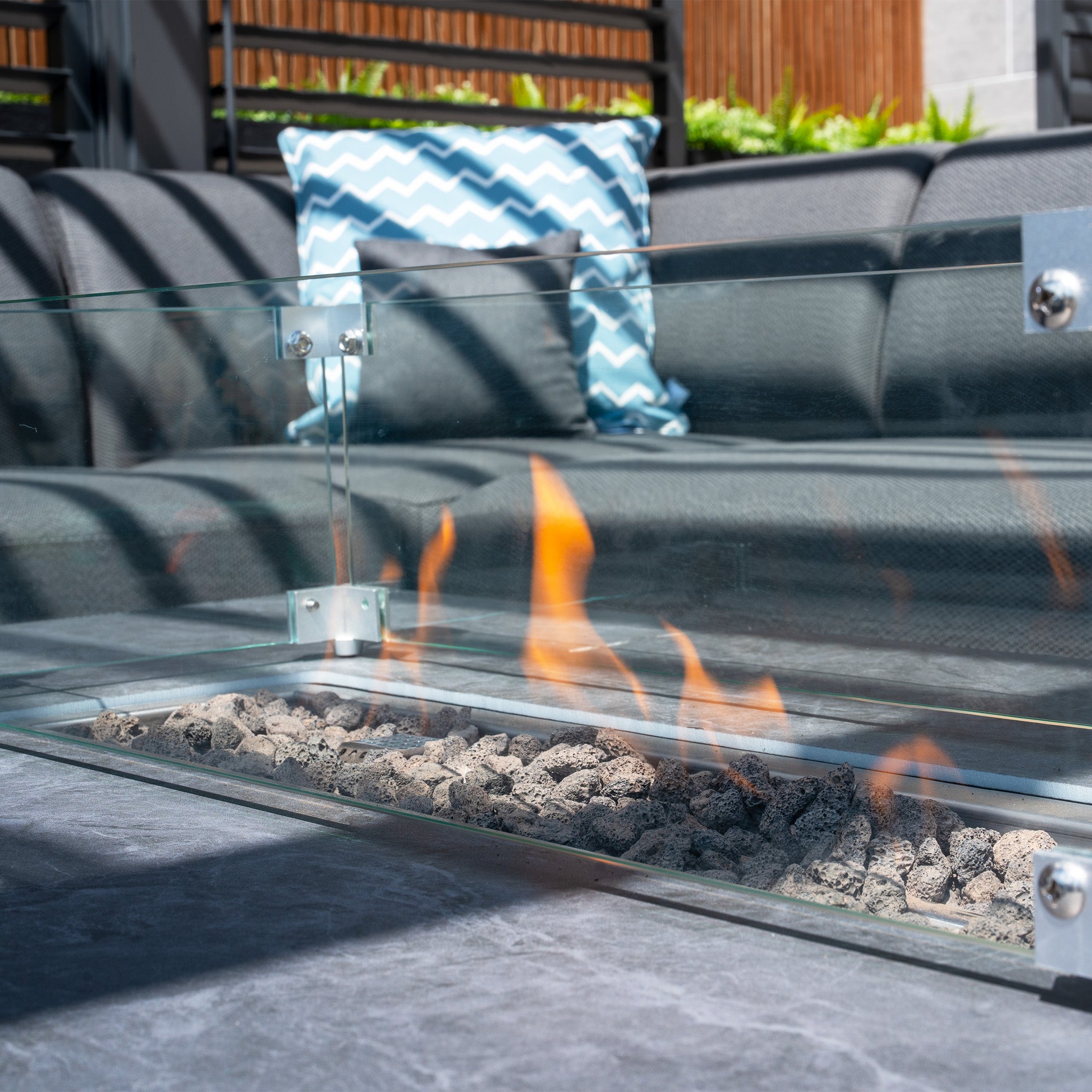 A Luna Outdoor Fabric Large Corner Group Set with Firepit Coffee Table in Grey, featuring a glass enclosure, enhances the patio, complemented by a chevron-patterned pillow.