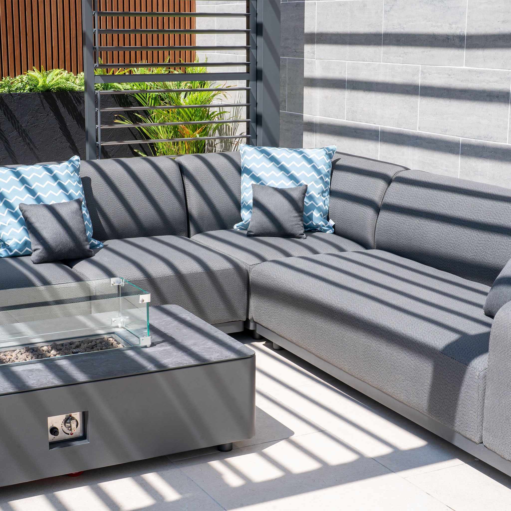 The Luna Outdoor Fabric Large Corner Group Set with Firepit Coffee Table in Grey includes a gray sectional sofa with blue wave-patterned cushions and a sleek firepit coffee table, all set under stylish striped shades.