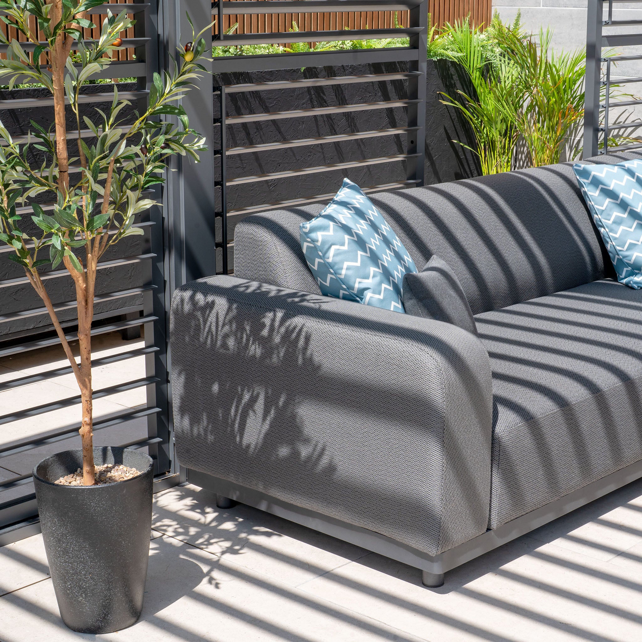 The Luna Outdoor Fabric Large Corner Group Set in Grey, paired with a firepit coffee table and two blue patterned pillows, offers the ultimate stylish relaxation under slatted shade with plant surroundings.