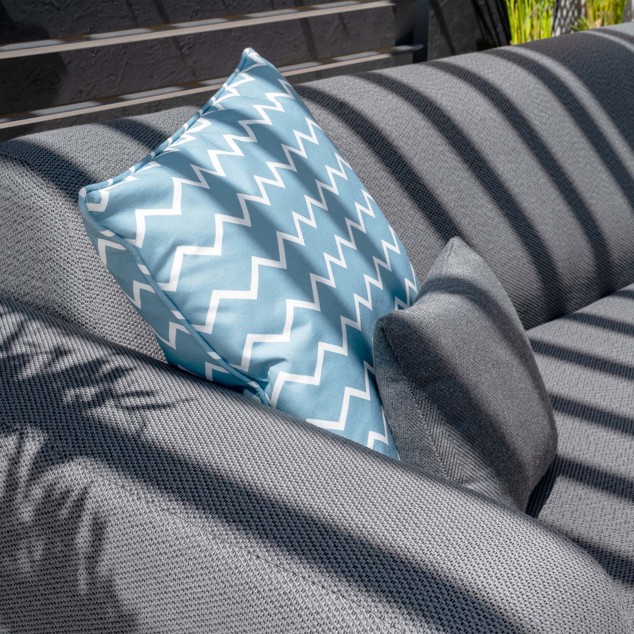 A gray sofa with a blue and white zigzag-patterned pillow and a smaller gray pillow is bathed in sunlight, reflecting the stylish comfort of the Luna Outdoor Fabric Large Corner Group Set with Firepit Coffee Table in Grey, ideal for an inviting outdoor space.