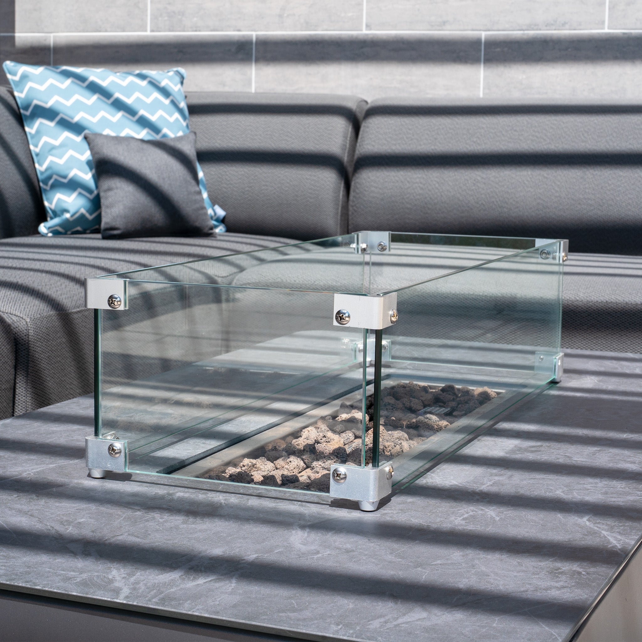 The Luna Outdoor Fabric Large Corner Group Set with a gray firepit coffee table, glass tabletop fire pit with black stones, and a gray sofa with blue patterned pillows beautifully completes any outdoor space.