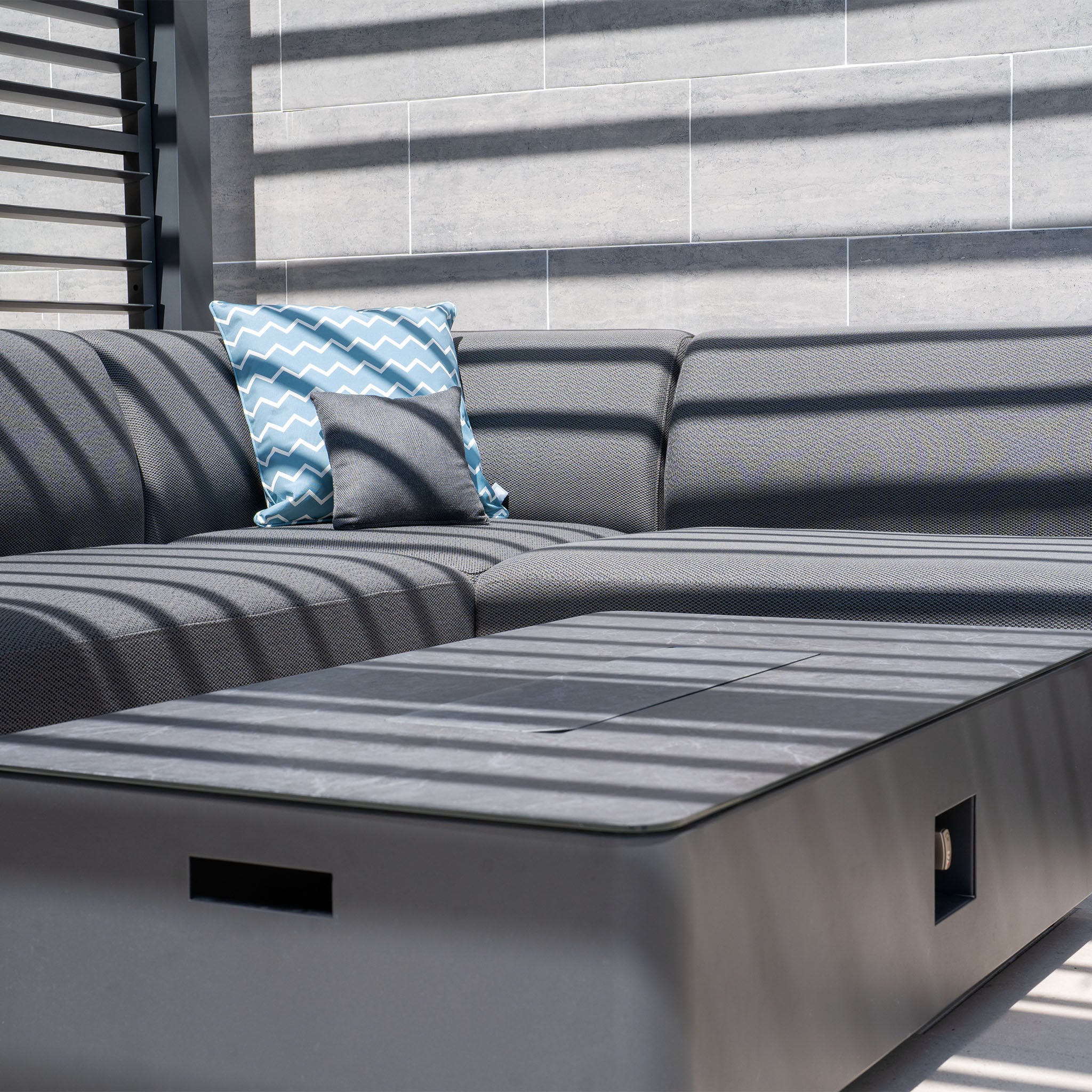 The Luna Outdoor Fabric Large Corner Group Set in Grey, adorned with a chevron-patterned pillow and illuminated by sunlight, features shadow stripes from the window. This cozy ambiance is enhanced by the complementary Firepit Coffee Table, perfect for any Harbour Lifestyle setting.
