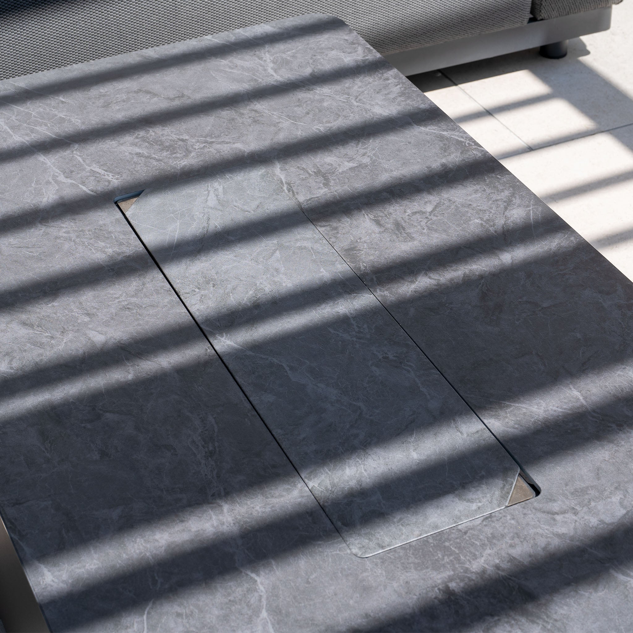 The Luna Outdoor Fabric Large Corner Group Set with Firepit Coffee Table in Grey features a gray stone surface table with a rectangular cutout, enhanced by slatted shadows. Its minimalistic design pairs perfectly for a chic yet cozy outdoor style.