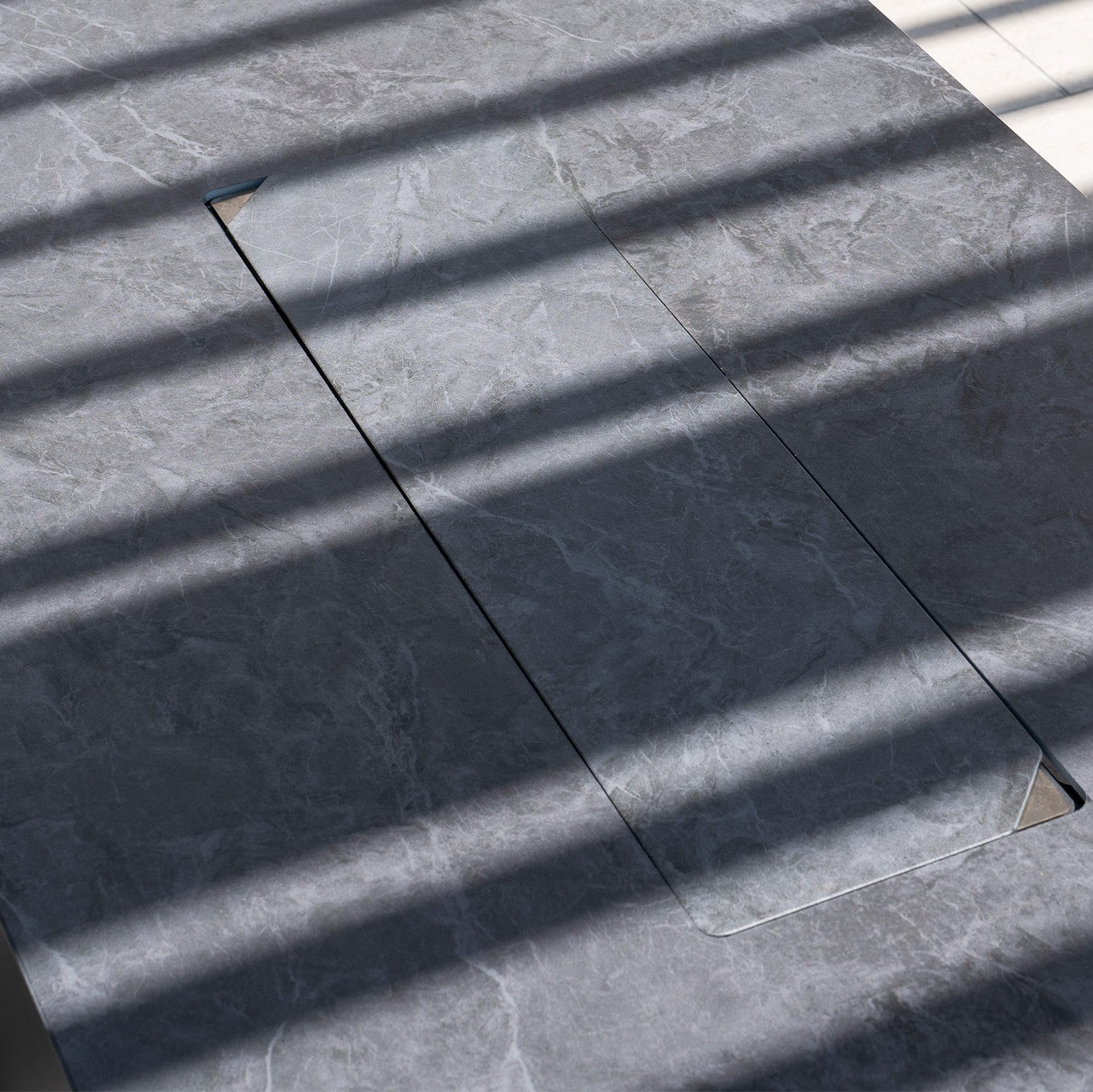 Diagonal shadows cross a gray marble surface with a rectangular cutout—perfect inspiration for the Luna Outdoor Fabric Corner Sofa Set with Firepit Coffee Table in Fawn, ideal for complementing your outdoor space.