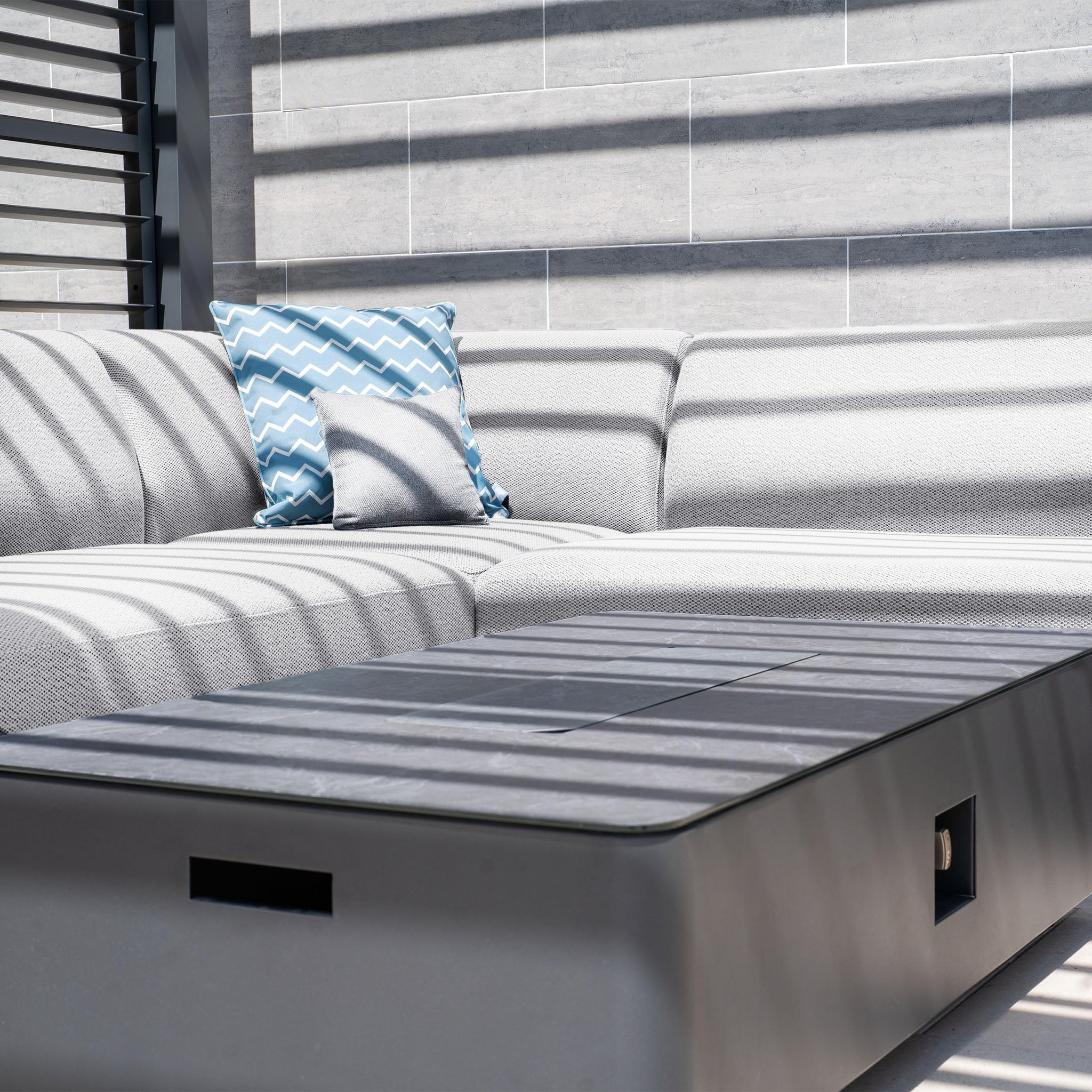 An outdoor sofa set in stain-proof Luna Outdoor Fabric, featuring a blue zigzag pillow and sleek firepit coffee table under striped shadows from a slatted roof.