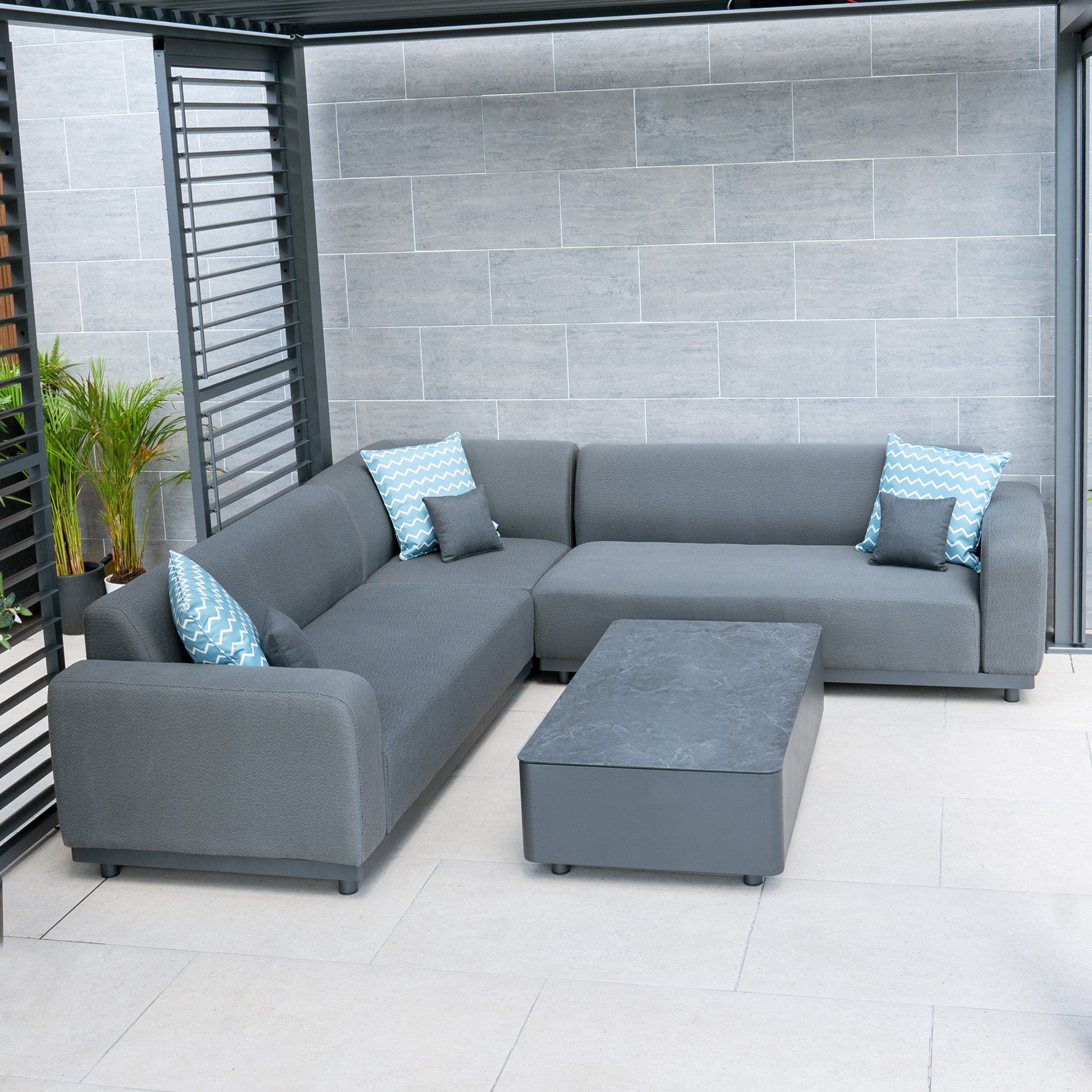 A modern outdoor patio showcases the Luna Outdoor Fabric Corner Sofa Set with Coffee Table in Grey, featuring a stain-proof sectional sofa alongside blue-patterned pillows.