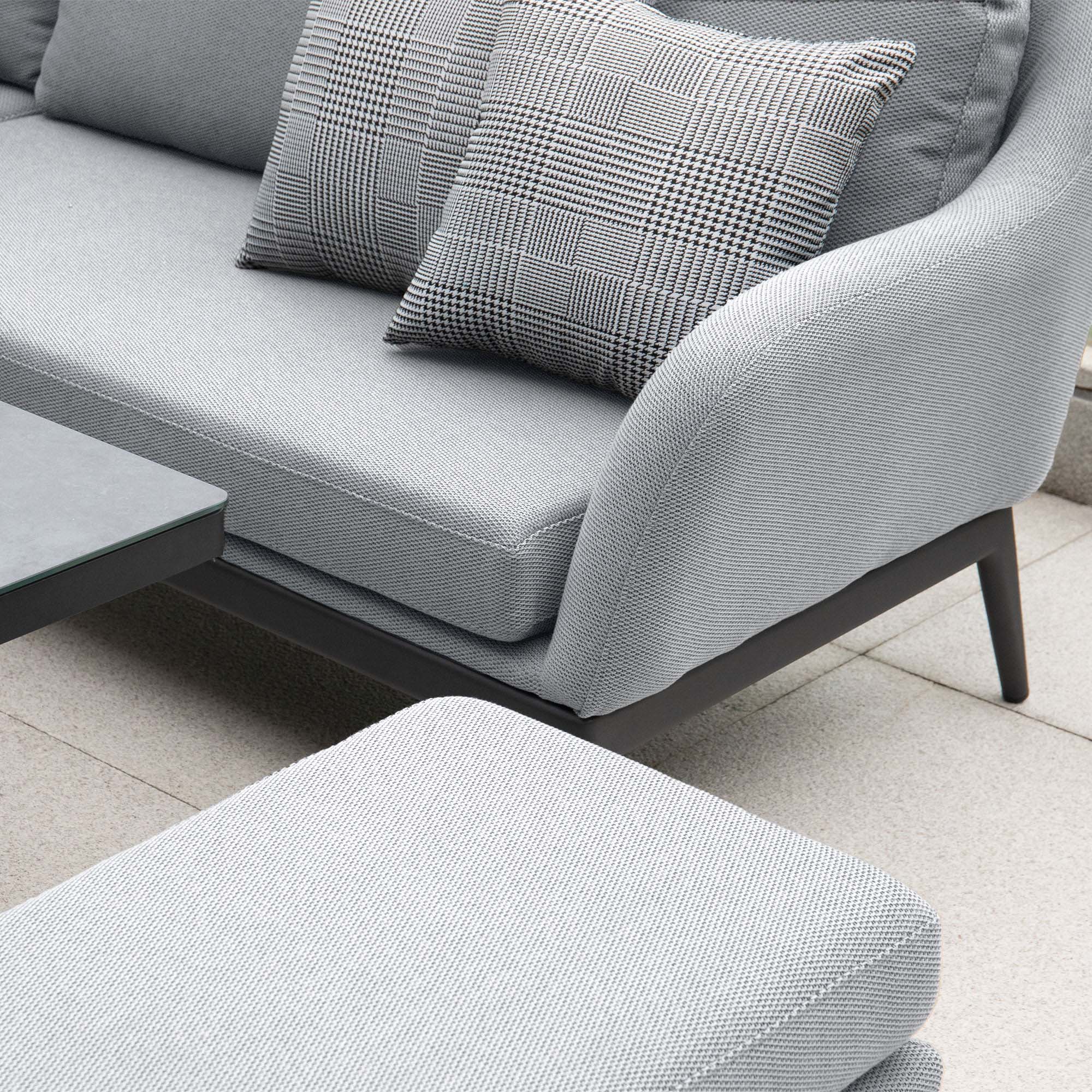 A modern gray outdoor sofa with checkered cushions and a matching ottoman on a stone tile patio, complemented perfectly by the Luna U-Shape Outdoor Fabric Corner Dining with Rising Table in Oyster Grey.