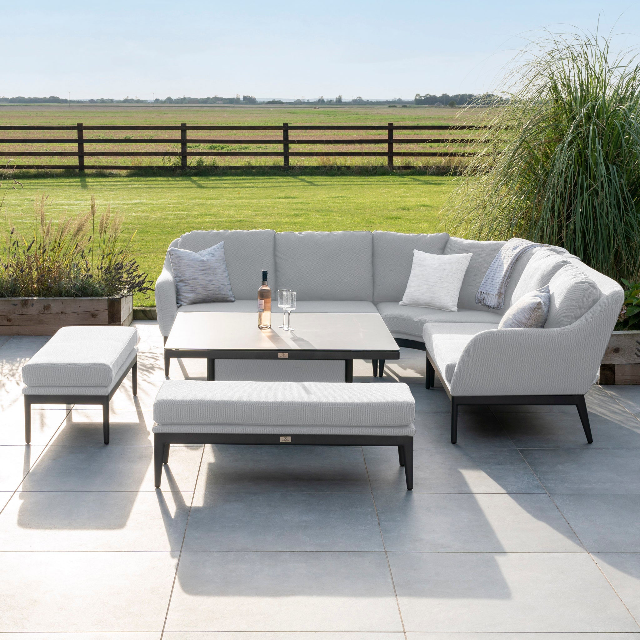 Luna Deluxe Outdoor Fabric Square Corner Dining Set in Oyster Grey, featuring a cushioned gray sectional sofa, two benches, and a maintenance-free rectangular coffee table with a wine bottle on the table.