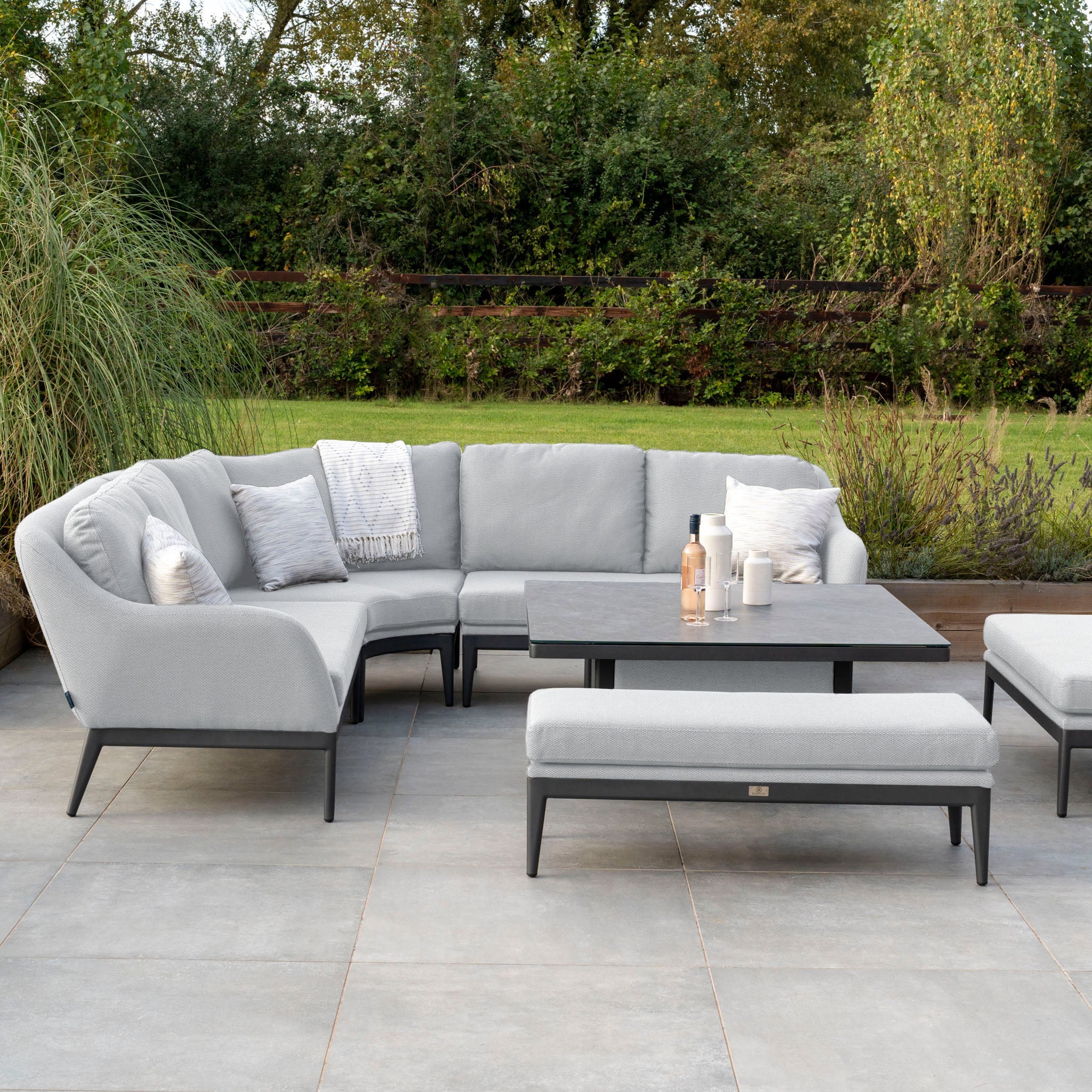 Outdoor patio featuring the Luna Deluxe Outdoor Fabric Square Corner Dining Set with a Rising Table in Oyster Grey, complemented by a matching bench, lush greenery, and pillows. Enjoy relaxation with Luna outdoor fabric cushions designed for a maintenance-free experience.