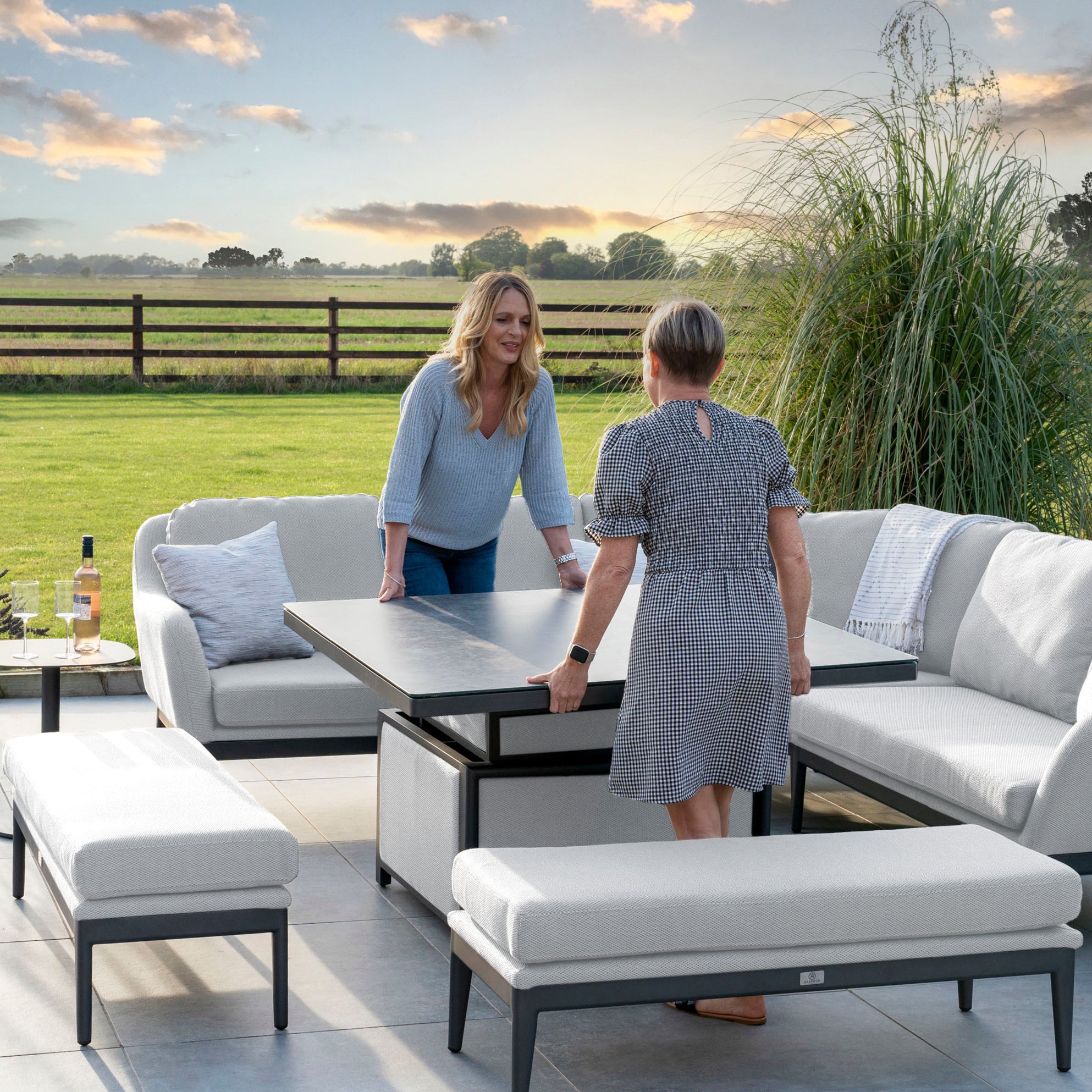Two women adjusting the Rising Table from the Luna Deluxe Outdoor Fabric Square Corner Dining Set in Oyster Grey on a patio with outdoor seating, overlooking a scenic field and wooden fence at sunset.