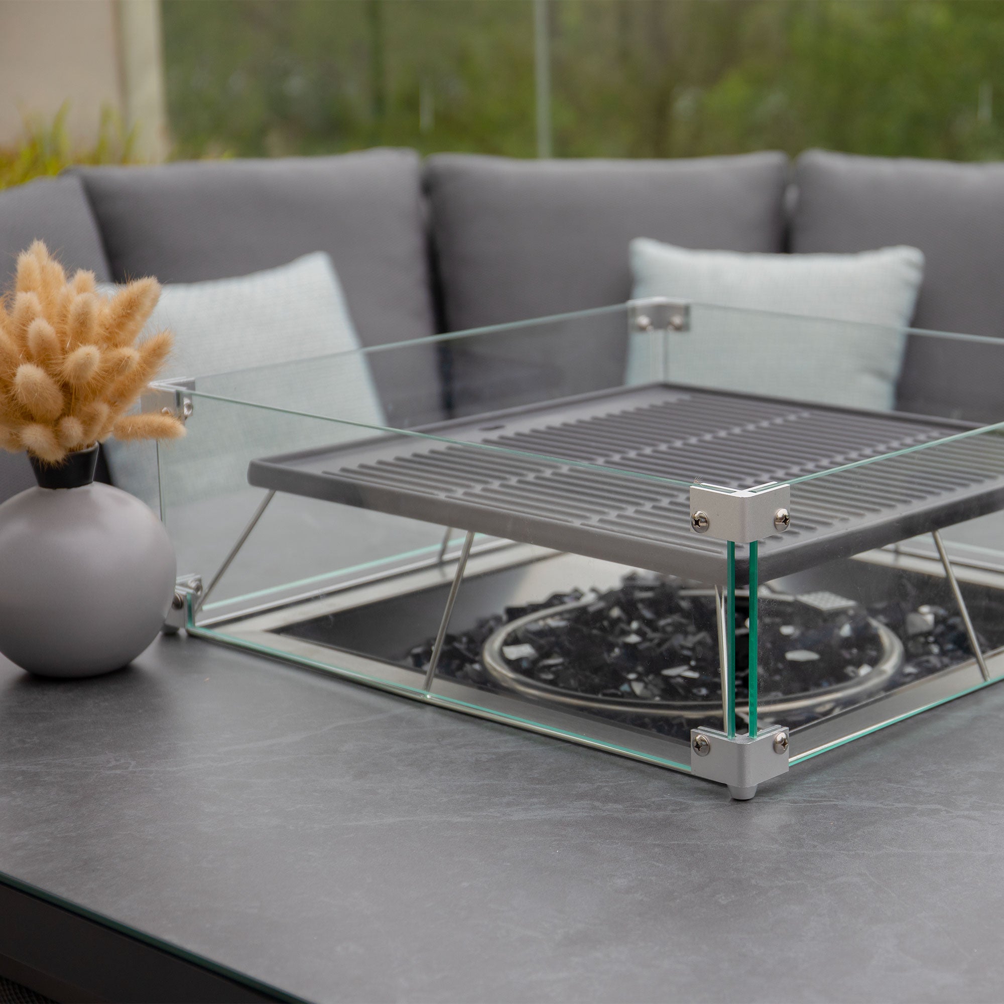 A rising firepit table sits on a patio, accompanied by a vase of beige dried flowers and an elegant Luna U-Shape Outdoor Fabric Corner Dining set in Grey in the background.