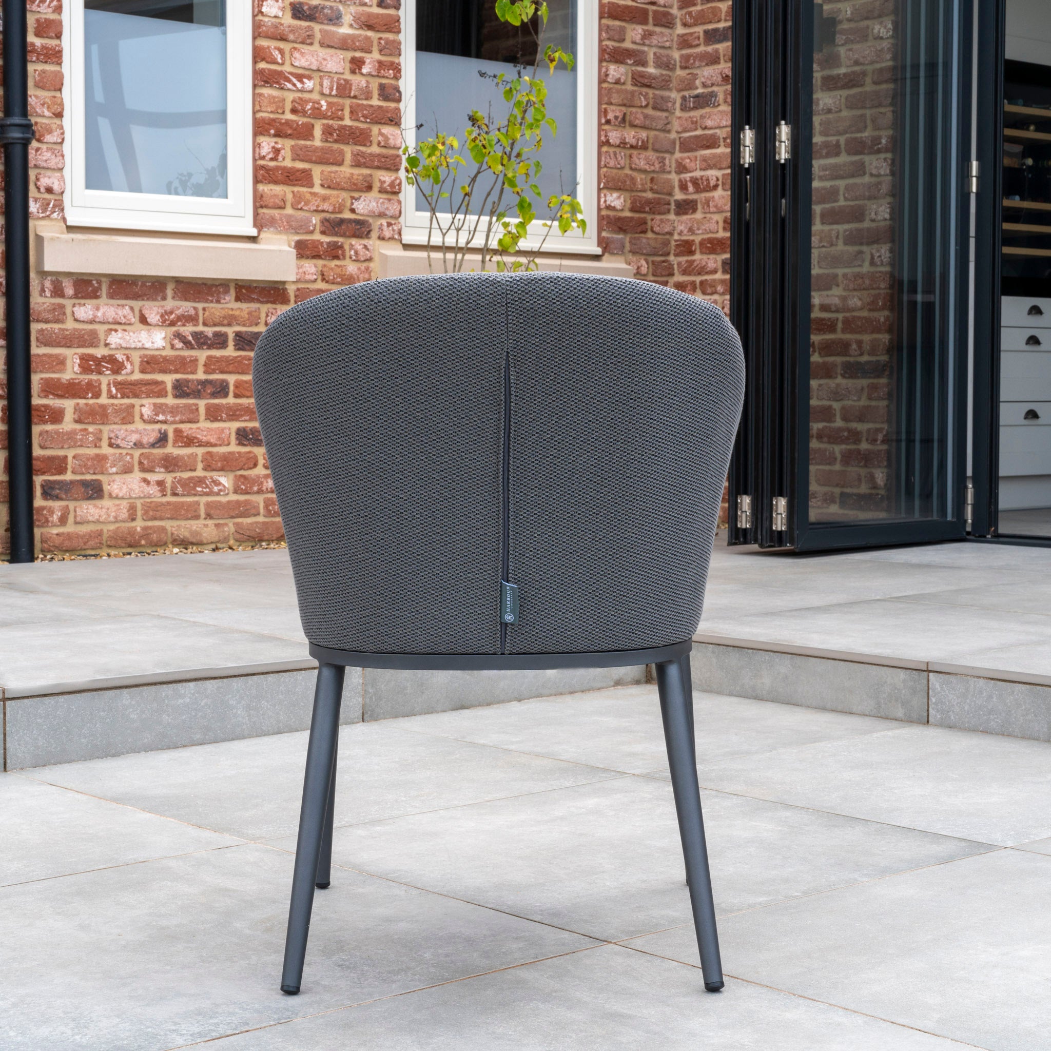 Part of the Luna 8 Seat Outdoor Fabric Extending Ceramic Dining Set in Grey, a stain-proof grey chair sits on a tiled patio in front of a brick house with glass doors, adorned by a potted plant in the background.