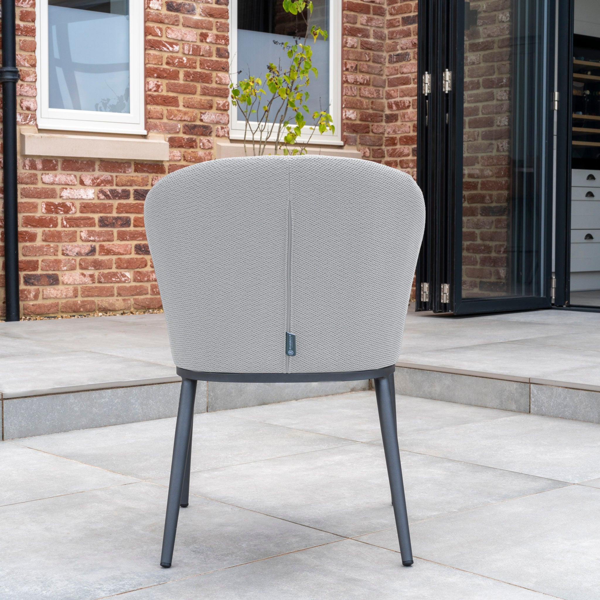 The Luna 8 Seat Outdoor Fabric Extending Teak Dining Set in Oyster Grey, characterized by its light gray tone and black legs, adorns a patio framed by brick walls and large windows in the background.