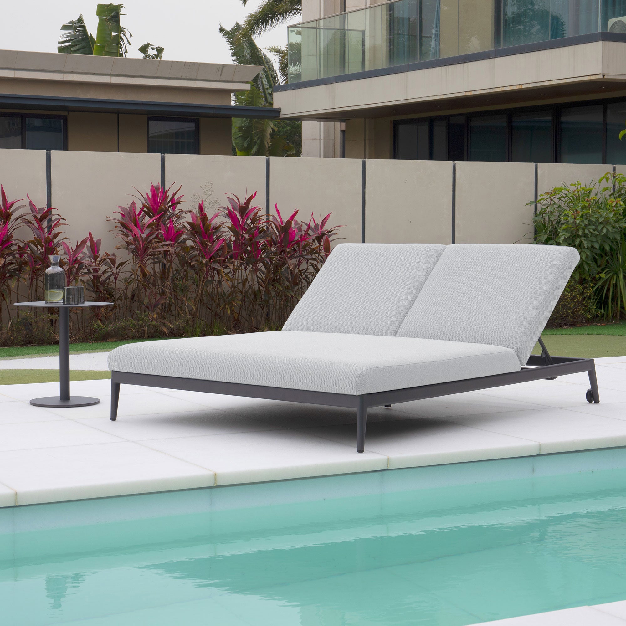 A stylish double sun lounger with Luna Outdoor Fabric in Oyster Grey, accompanied by two cushions, sits poolside next to a small round table holding a water bottle, offering both elegance and stain-proof durability.