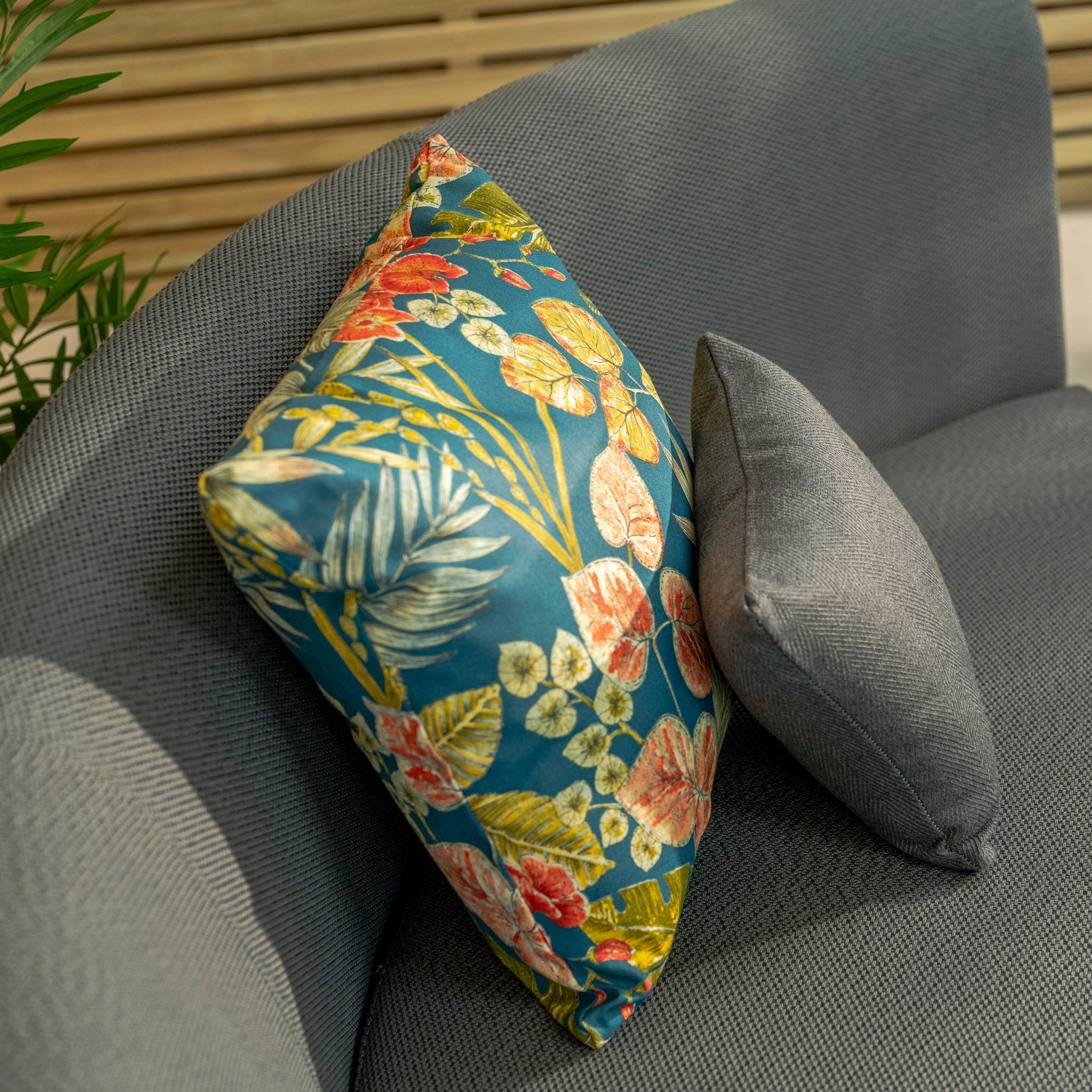 Two pillows adorn a grey couch from the "Luna Outdoor Fabric Lifestyle Suite with 2 Sofas and 2 Benches in Grey": one featuring a vibrant floral pattern and the other in plain grey, both made from Luna outdoor fabric. The stylish, maintenance-free setup is enhanced by wooden slats and a plant in the background.