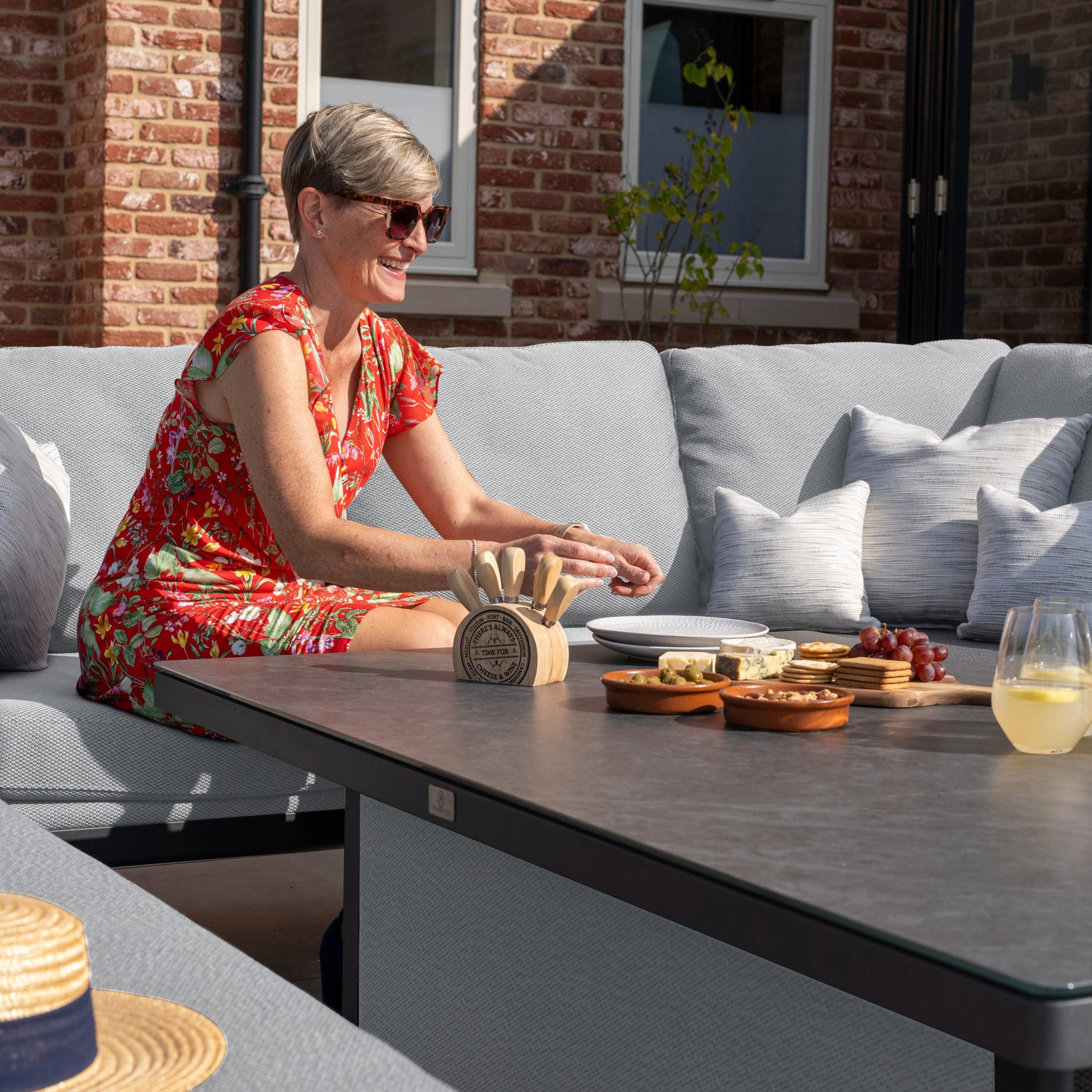 Woman in a red floral dress sitting on a patio sofa, preparing food on an outdoor table with various snacks and drinks, enjoying her Luna Outdoor Fabric Rectangular Corner Dining Set with Rising Table in Oyster Grey (Left Hand).