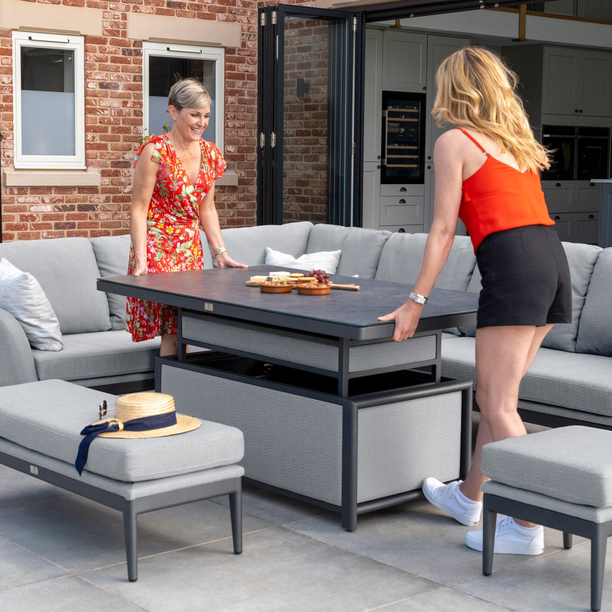 Two women are setting up a patio table with snacks, surrounded by the Luna Outdoor Fabric Rectangular Corner Dining Set with Rising Table in Oyster Grey (Left Hand), all against a charming brick wall.