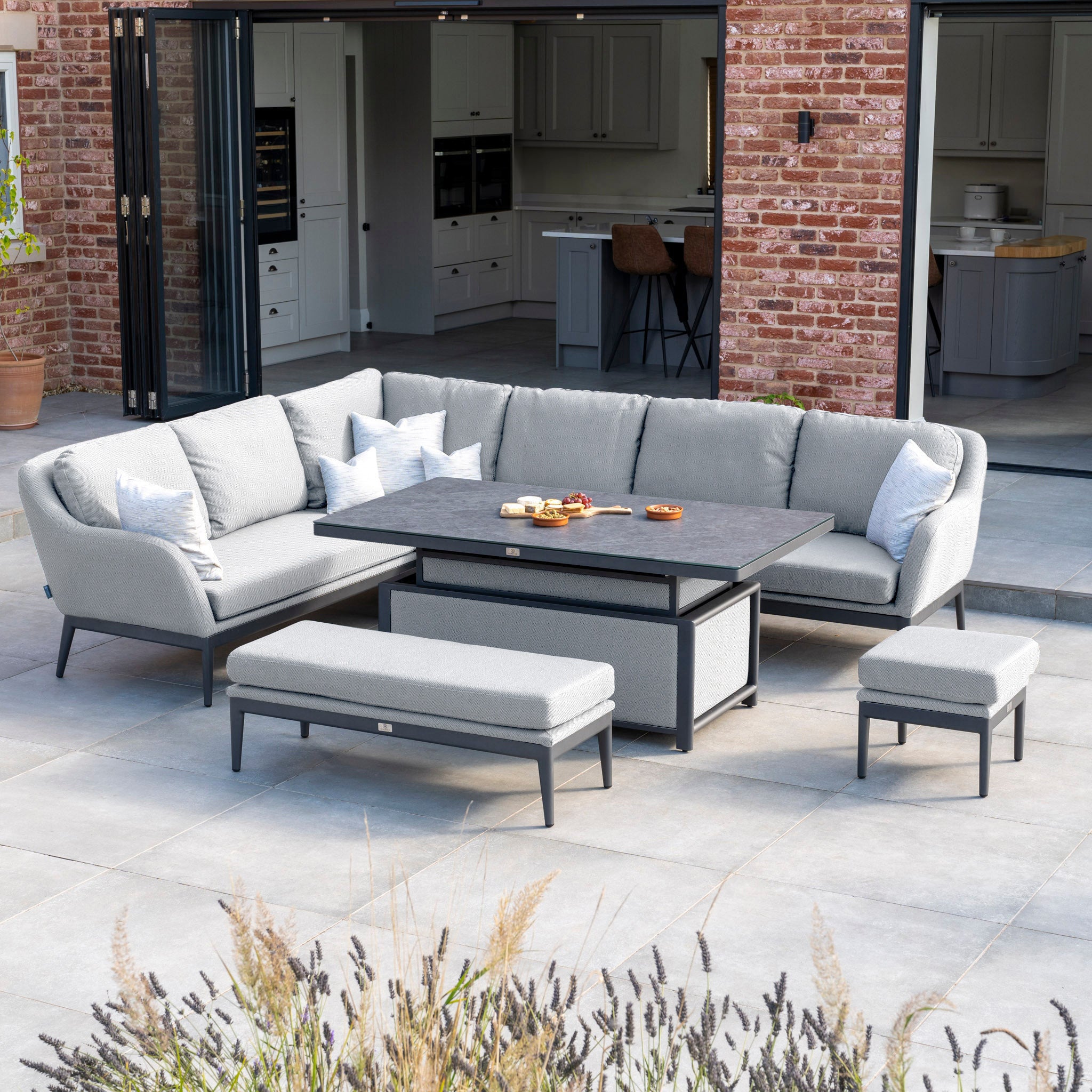 Outdoor patio featuring the Luna Outdoor Fabric Rectangular Corner Dining Set with Rising Table in Oyster Grey (Left Hand). This setup includes a gray sectional sofa, cushioned benches, and a black rectangular table near a brick house with open doors. The use of Luna outdoor fabric ensures lasting comfort and style, making it both chic and maintenance-free.
